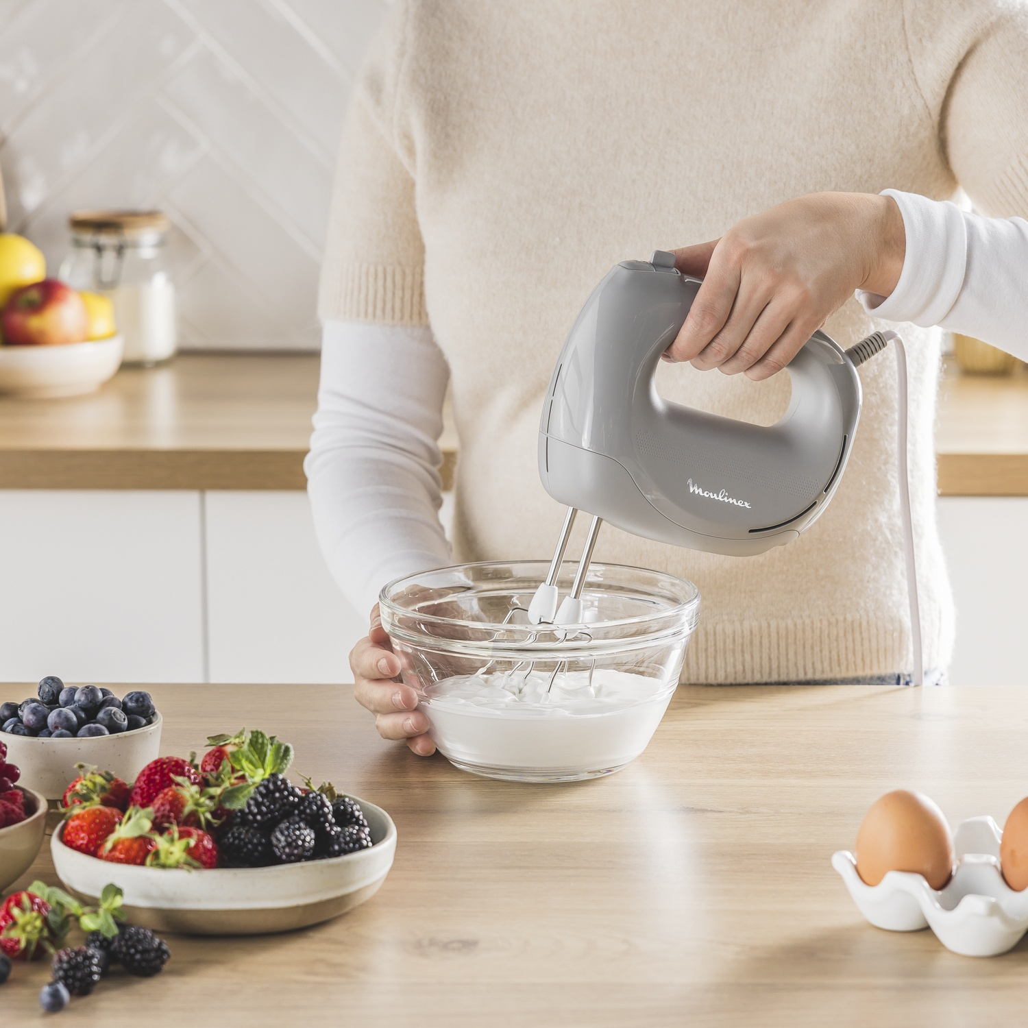 Een persoon gebruikt een grijze handmixer om slagroom op te kloppen in een glazen kom. Bessen en eieren liggen op tafel.