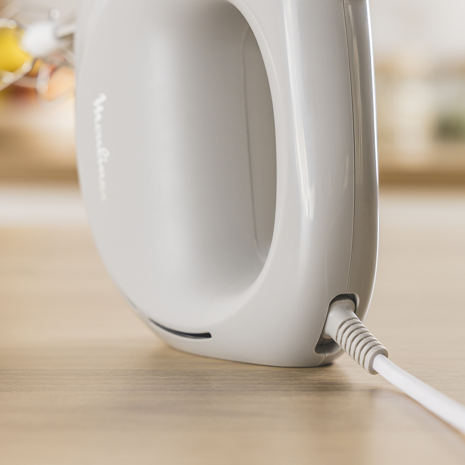 Close-up van een witte handmixer op een houten oppervlak. Het snoer van de mixer is aangesloten. De achtergrond is wazig.