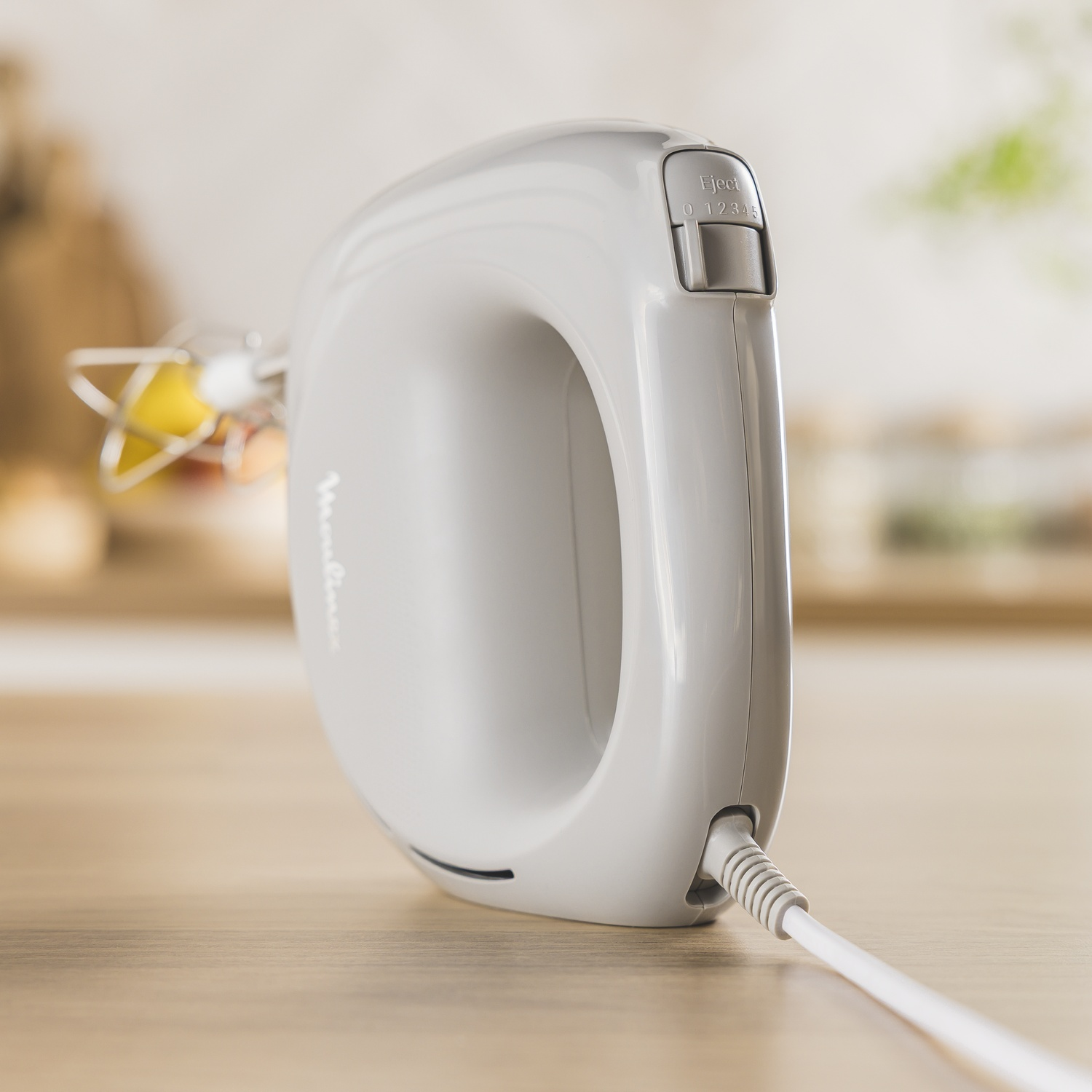 Een witte Moulinex handmixer op een houten oppervlak. De mixer heeft een snelheidsregelaar en een uitwerpknop. Een draad loopt uit de mixer.