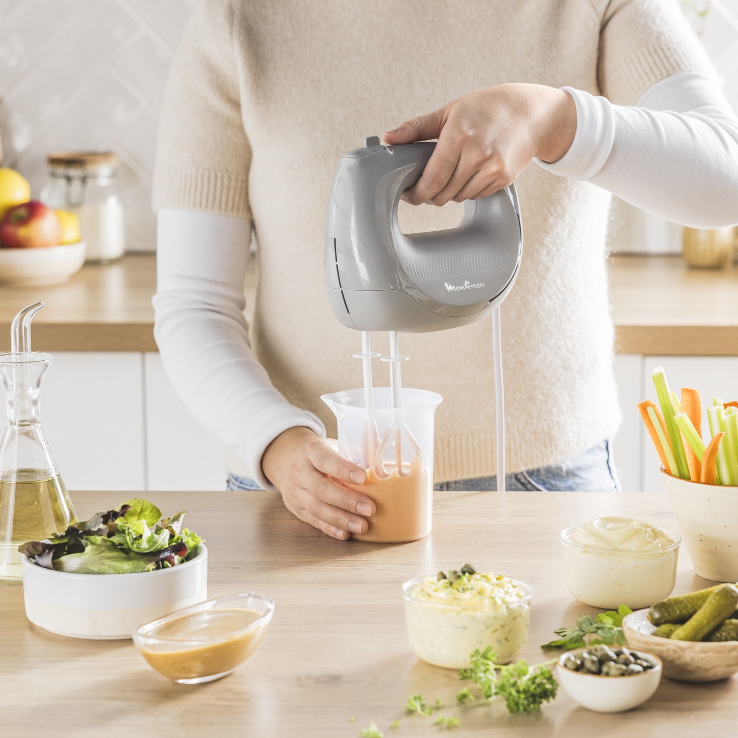 Een persoon gebruikt een handmixer om ingrediënten te mengen in een maatbeker. Diverse salade-onderdelen en dips staan op een tafel.