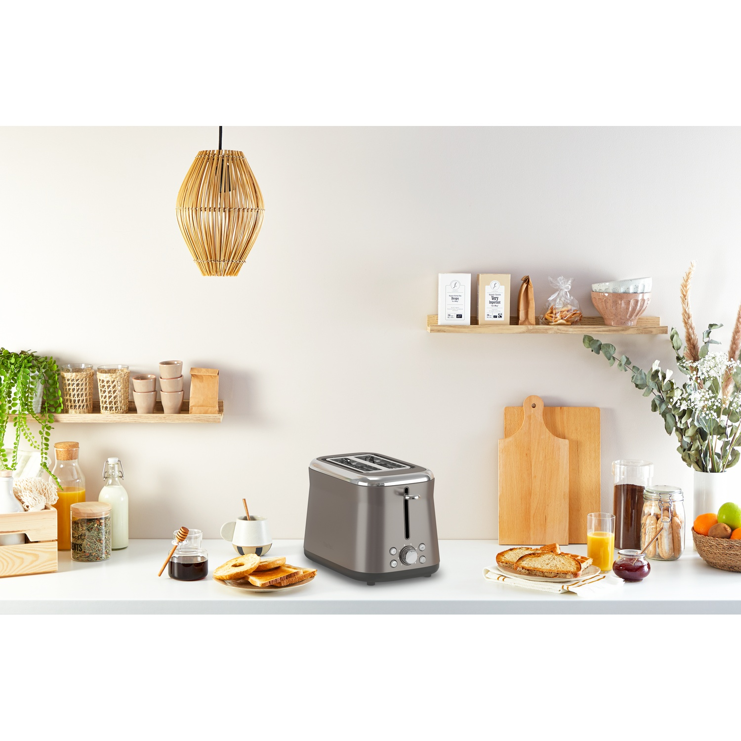A kitchen scene with a toaster, toast, honey, fruits, and plants. A woven lamp hangs above a white counter.