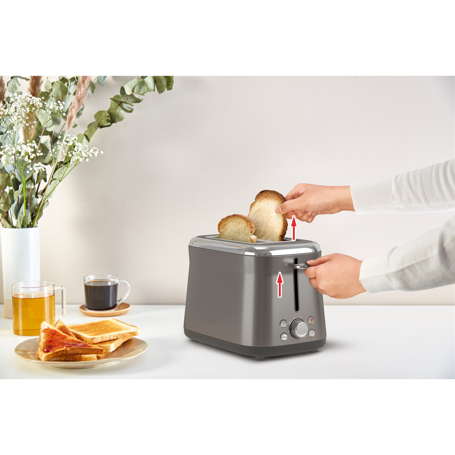 A person is taking out toasted bread from a gray toaster. On the table, there are slices of bread with jam, tea, and coffee.