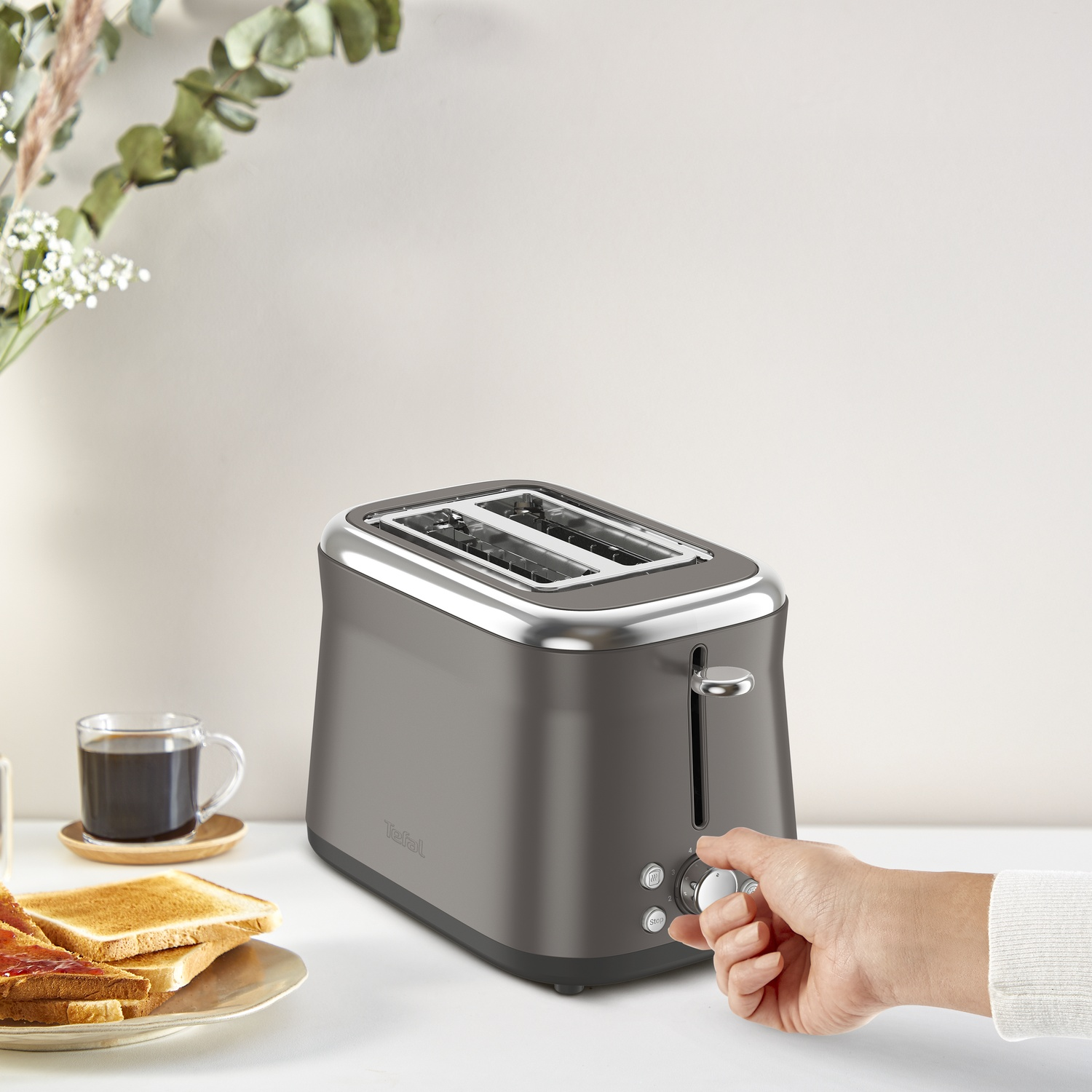 A gray toaster with a hand adjusting the settings. Toast and coffee are on the table next to the toaster.
