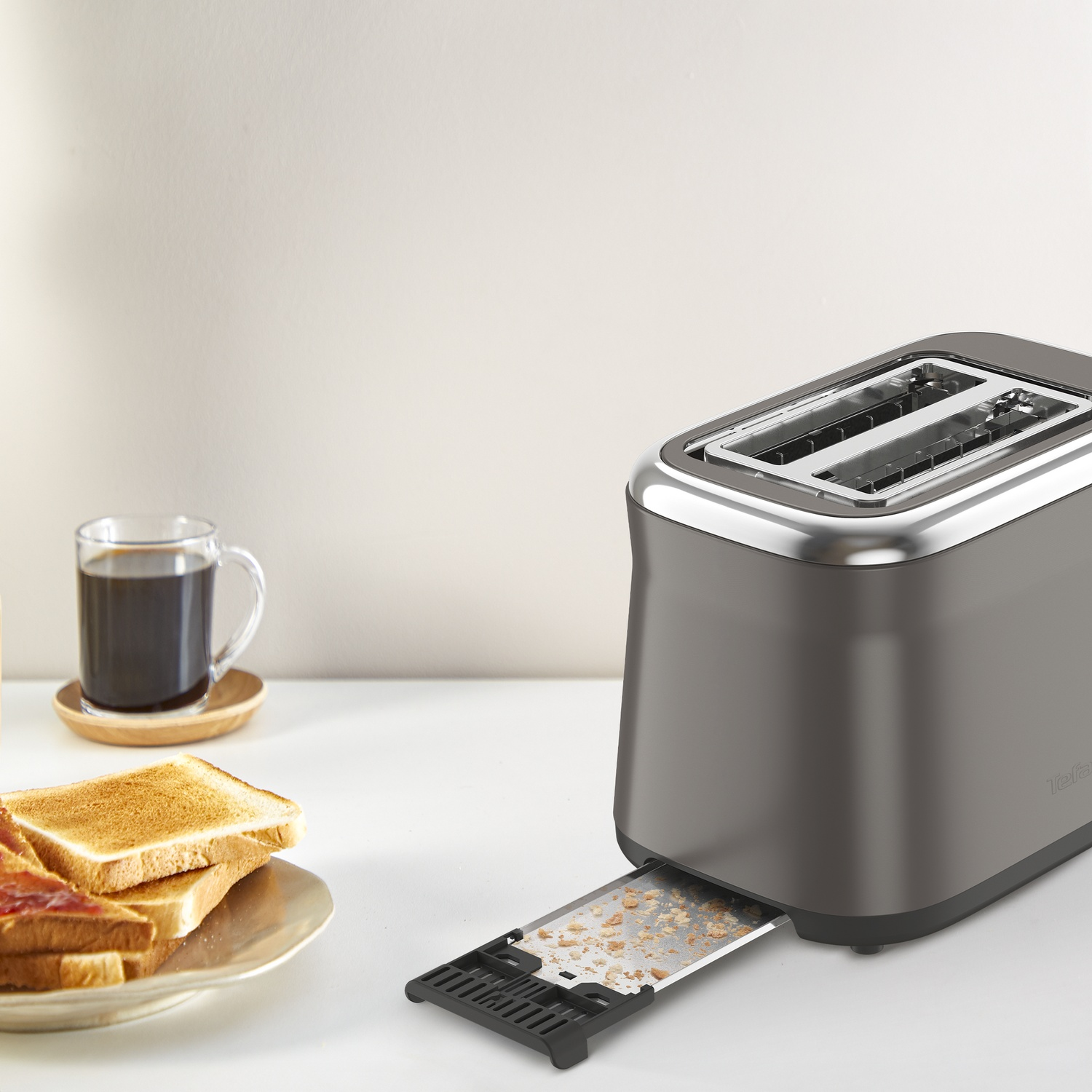 A toaster with toast and coffee. Crumbs are visible in the crumb tray. Toasted bread and jam on a plate.