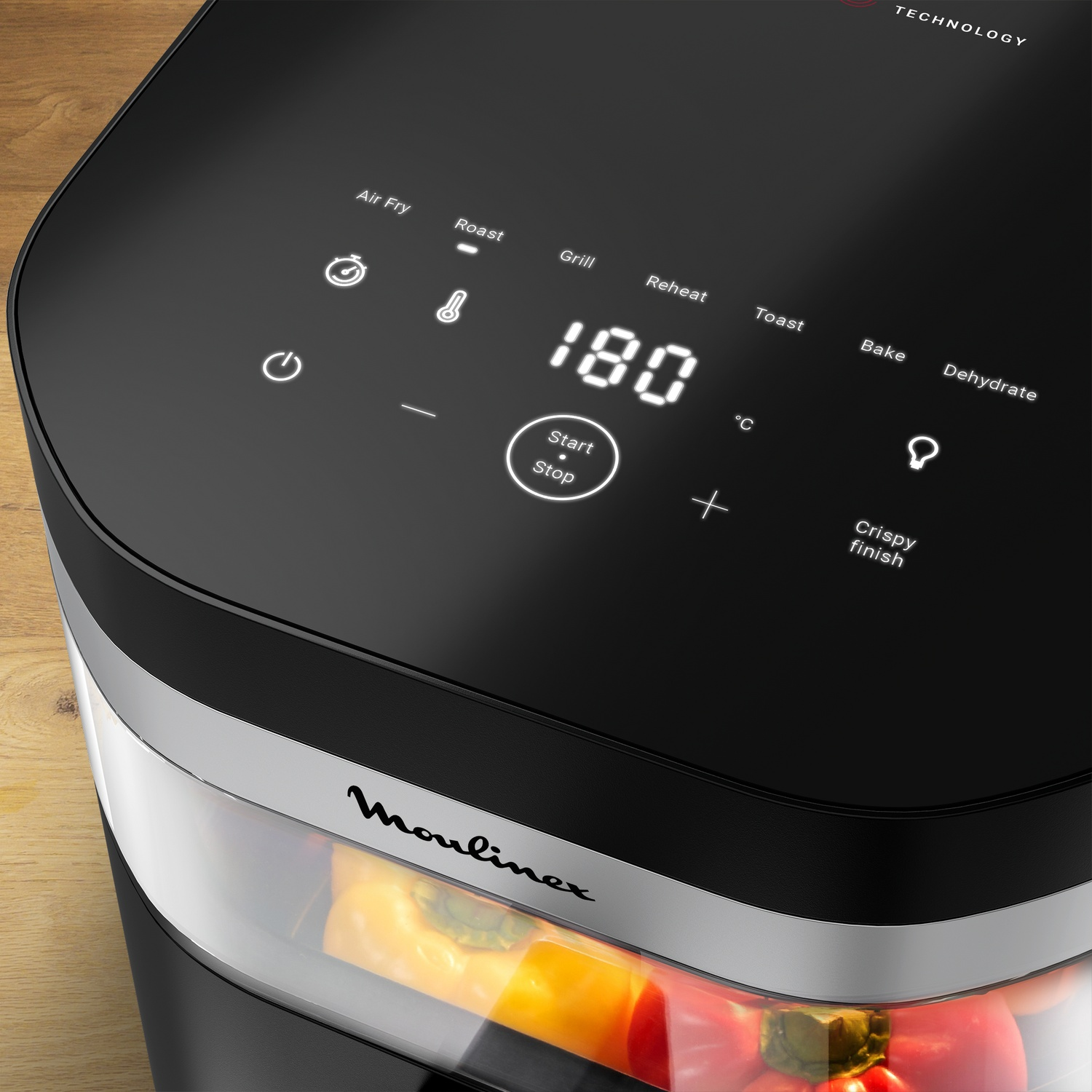 Close-up of a black air fryer with a digital display showing 180 degrees. The lower part of the fryer is transparent, showing colorful vegetables.