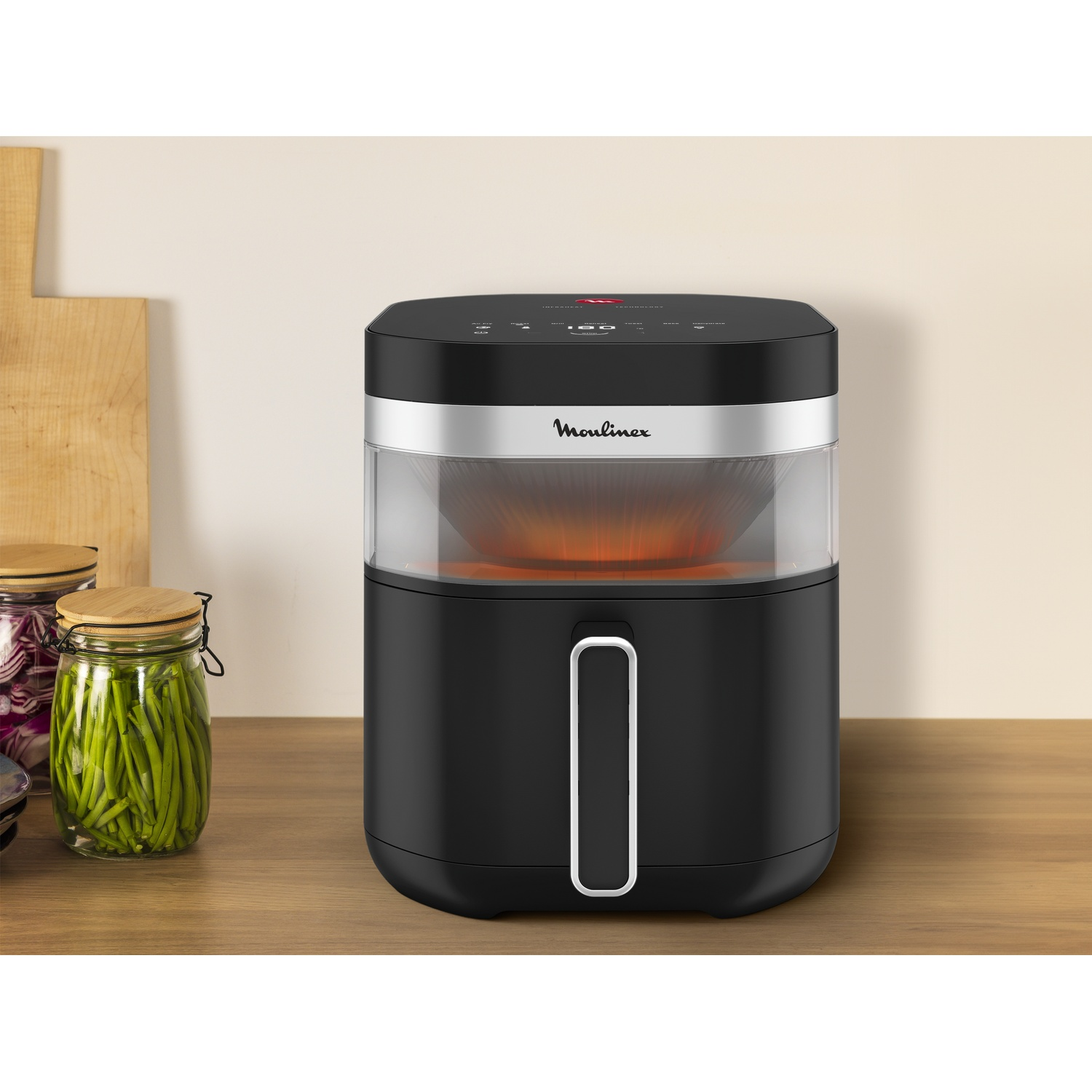 Black Moulinex air fryer on a wooden surface. Jars of vegetables and a cutting board are on the left. The fryer has a glass container.