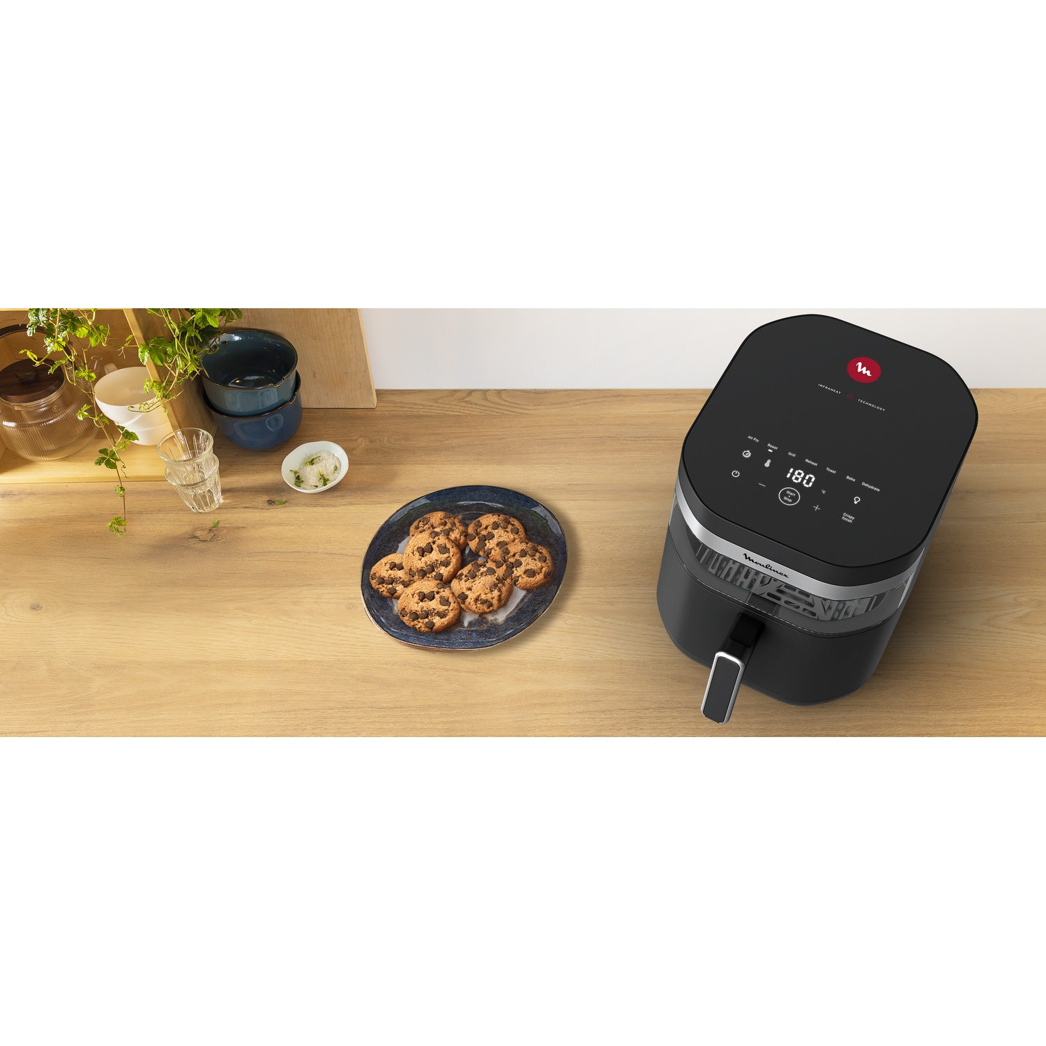 An air fryer and cookies on a wooden table. Other objects include bowls, a glass, and plants.