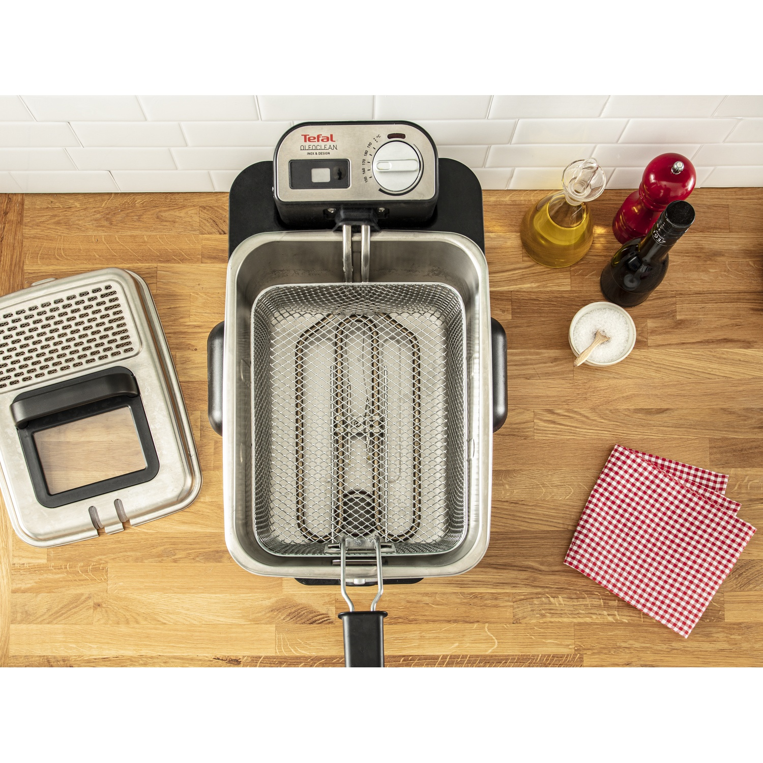 Vue aérienne d'une friteuse Tefal Oleoclean. De l'huile, du vinaigre, du sel et un tissu à carreaux rouges sont à proximité sur une surface en bois.