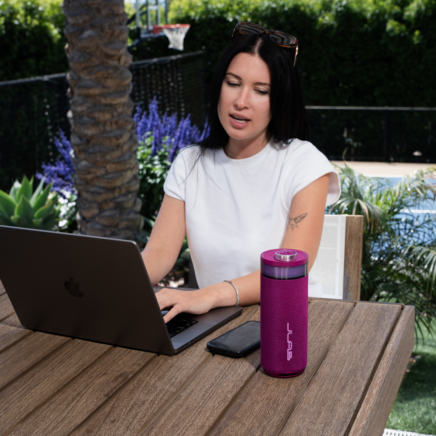Woman with laptop, sunglasses, and purple speaker sits at a wooden table outdoors. A phone is next to the speaker. Green background.