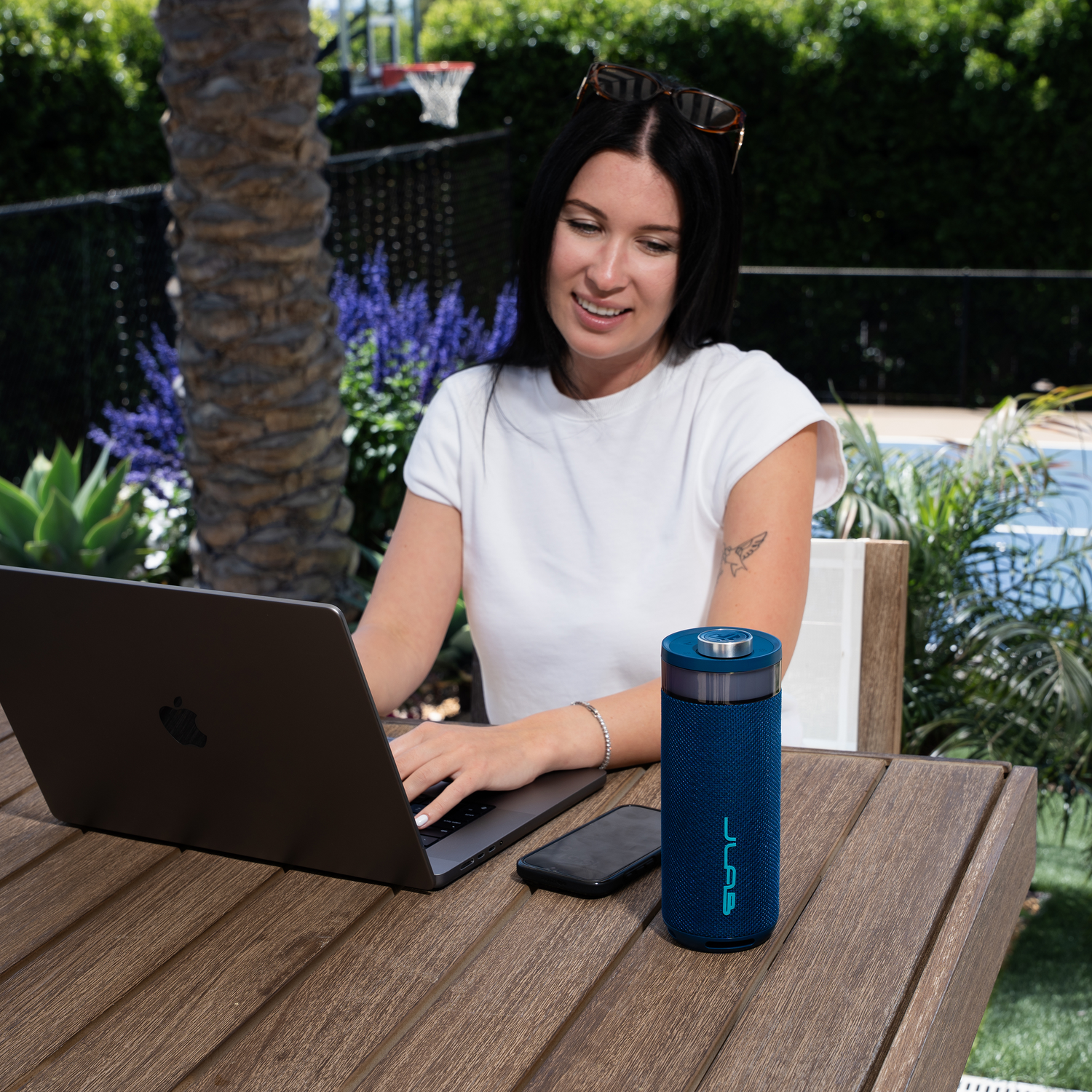 Woman uses a laptop outdoors, with a blue speaker and phone on the table. Sunglasses rest on her head.