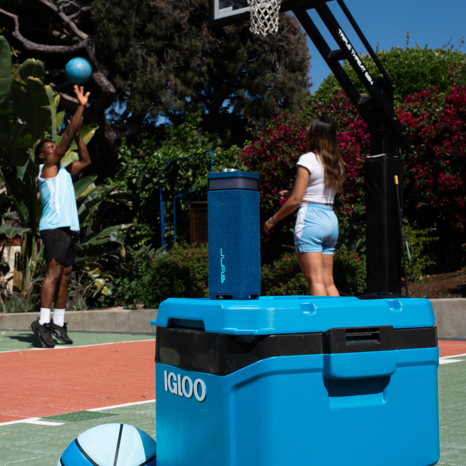 A person throws a basketball. A blue speaker is on a blue cooler. Another person stands near a basketball hoop.