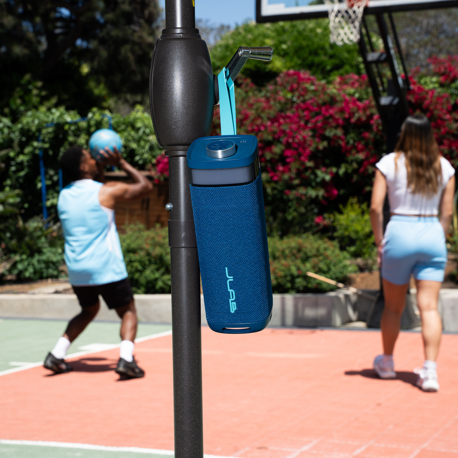 A blue speaker hangs from a basketball hoop. A person throws a ball, another walks towards the hoop. 
