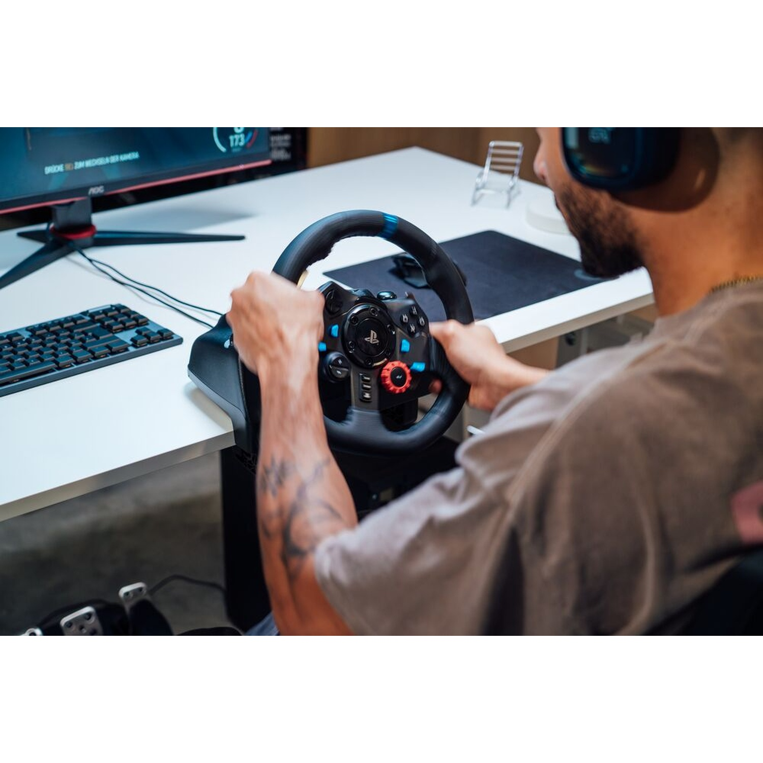 Una persona gioca a un gioco di corse con un volante. La scena include un monitor, una tastiera e pedali. La persona indossa le cuffie.