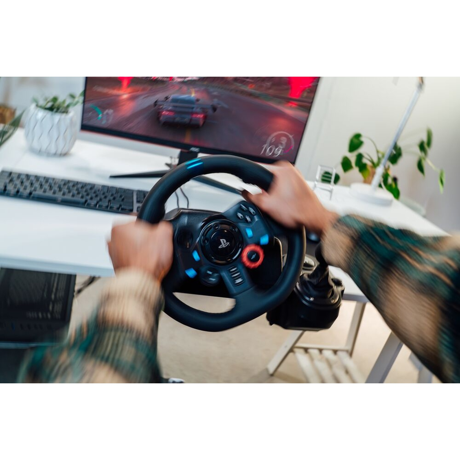 Mani su un volante, che giocano a un gioco di corse. Un monitor di computer mostra il gioco. Sono visibili anche una tastiera e delle piante.