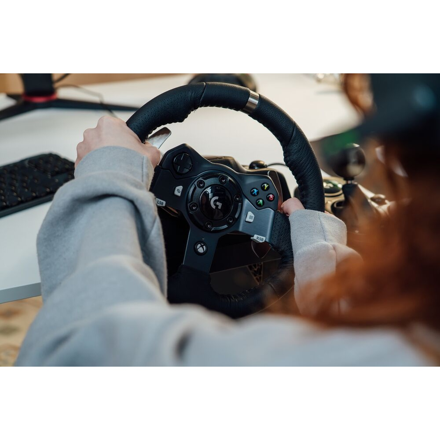 Mani che tengono un volante nero con pulsanti. Il volante è centrato e la persona indossa una felpa grigia.