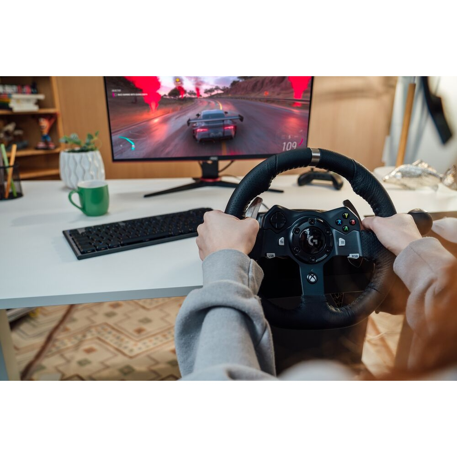 Mani che tengono un volante, che giocano a un gioco di corse su un monitor. Una tastiera e una tazza verde sono sulla scrivania.