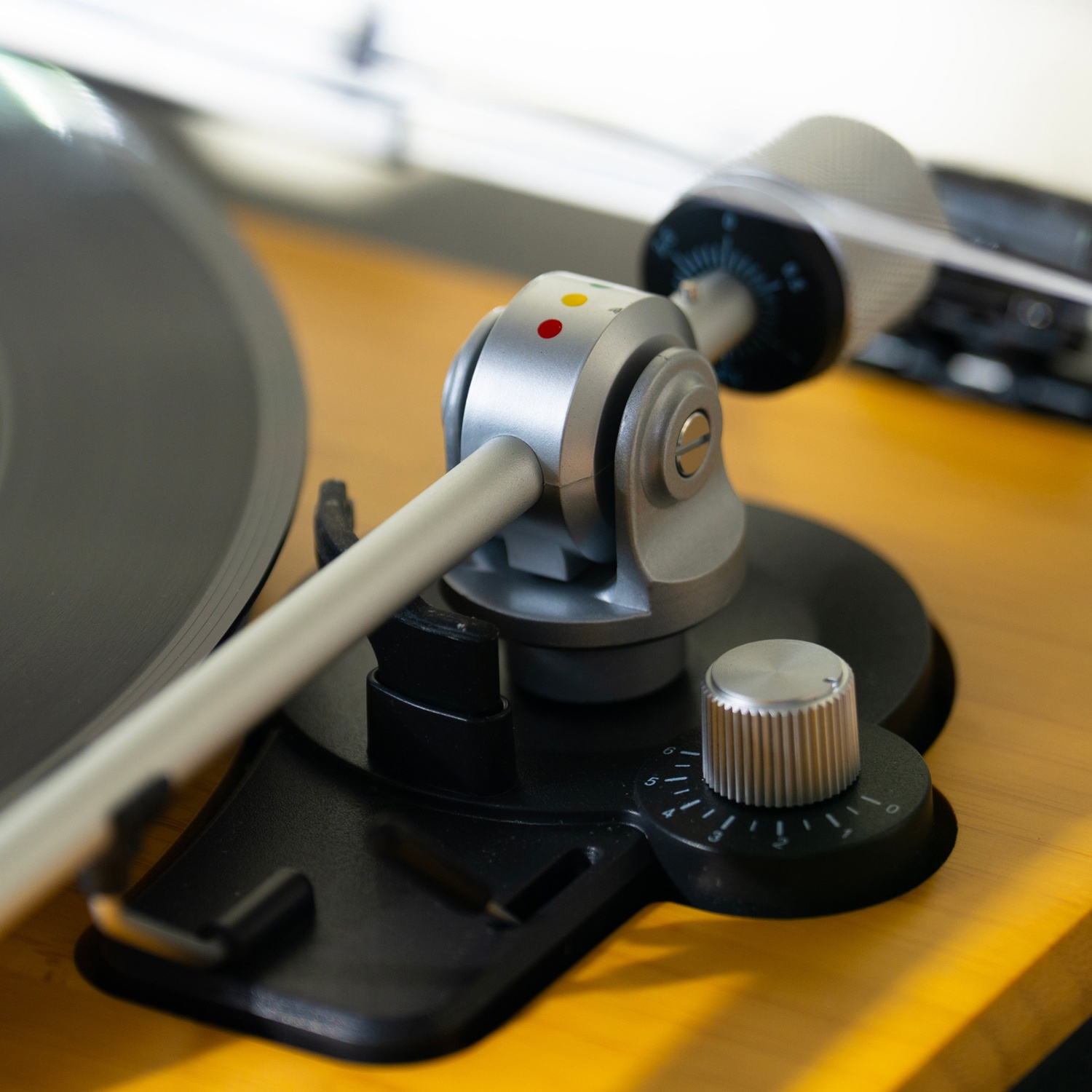 Gros plan sur le bras de lecture d'une platine. Un disque vinyle noir à gauche. Le bras est argenté avec un point rouge et jaune.