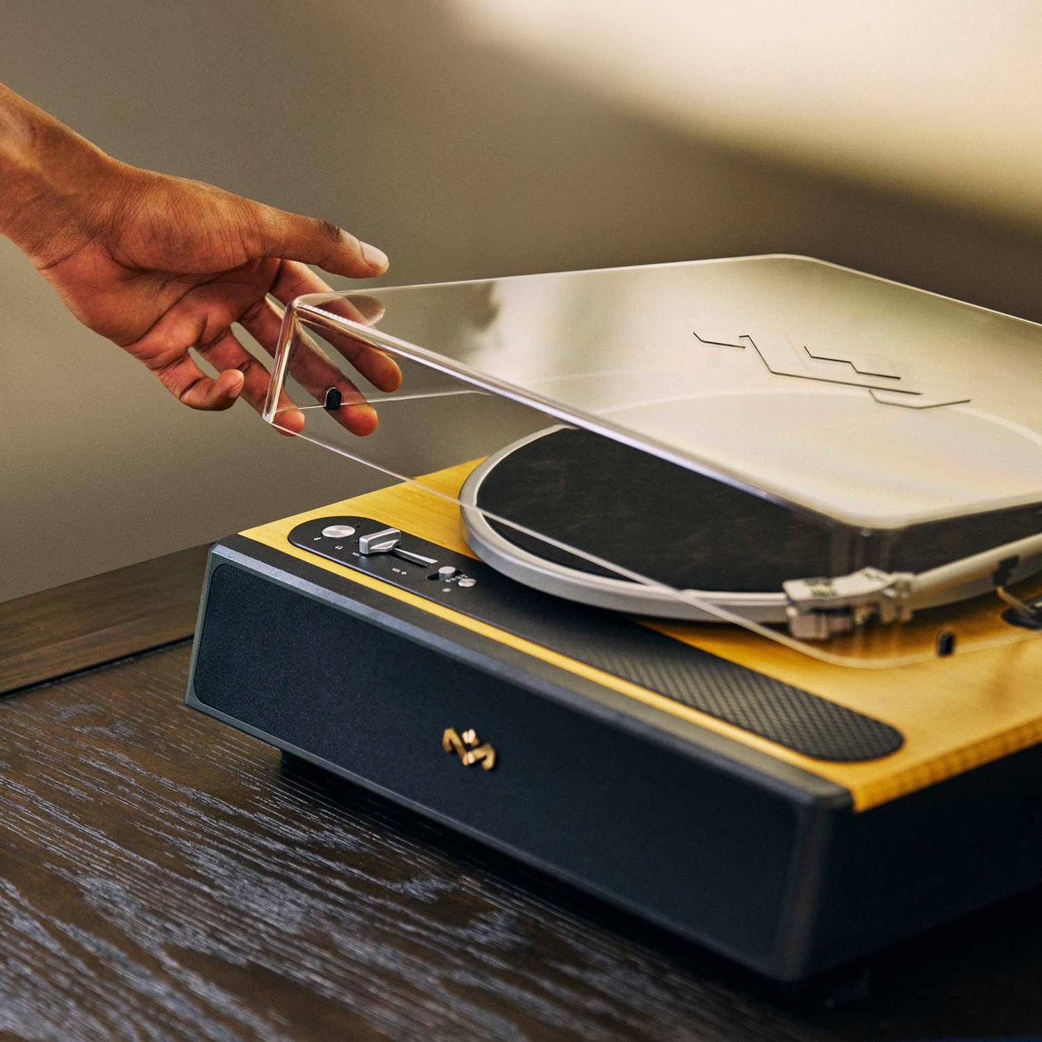 Une main soulève le couvercle transparent d'une platine vinyle en bois et noire. La platine est sur une surface en bois sombre.