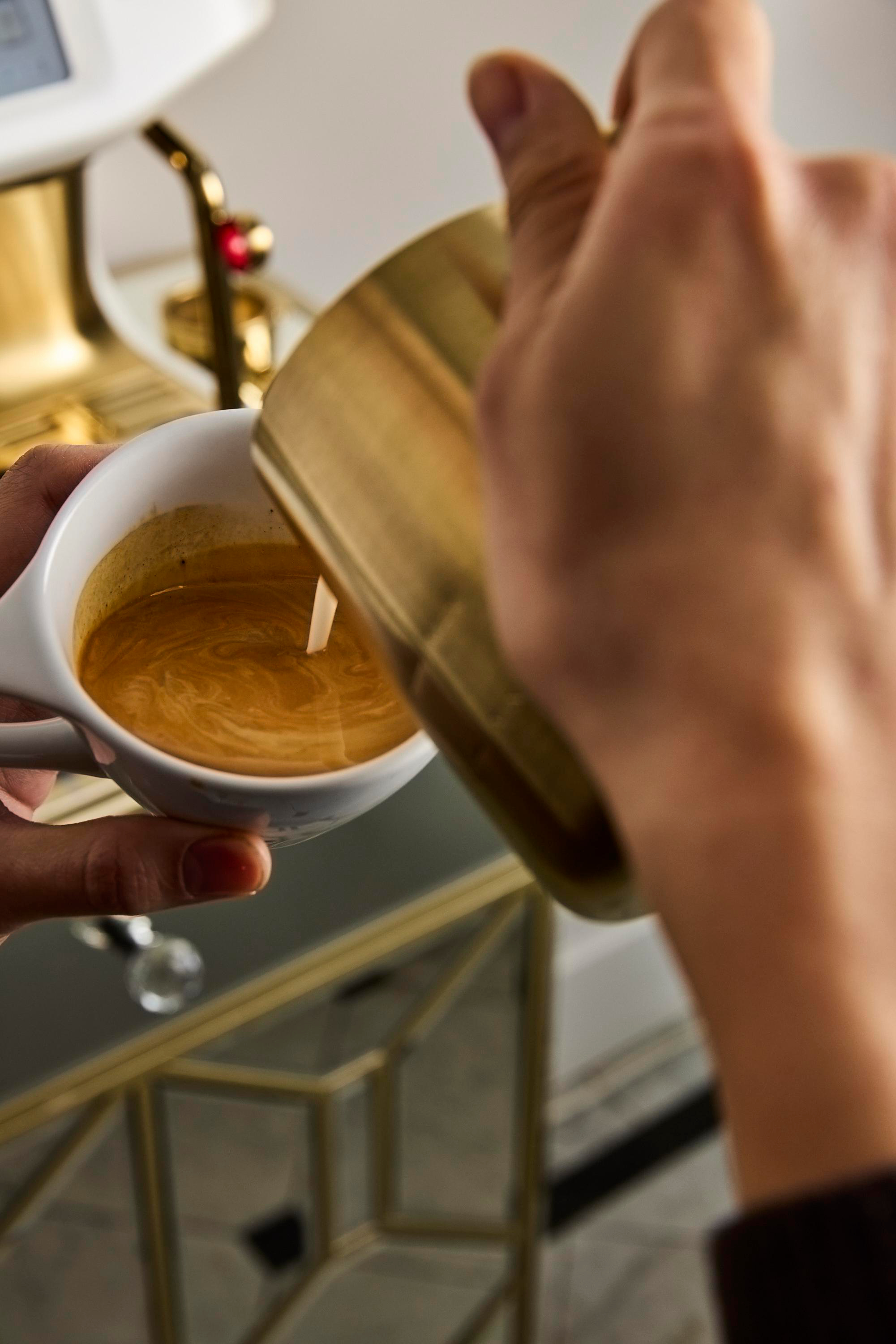 Hände gießen Milch aus einem goldenen Krug in eine Tasse Kaffee. Im Hintergrund eine Kaffeemaschine.