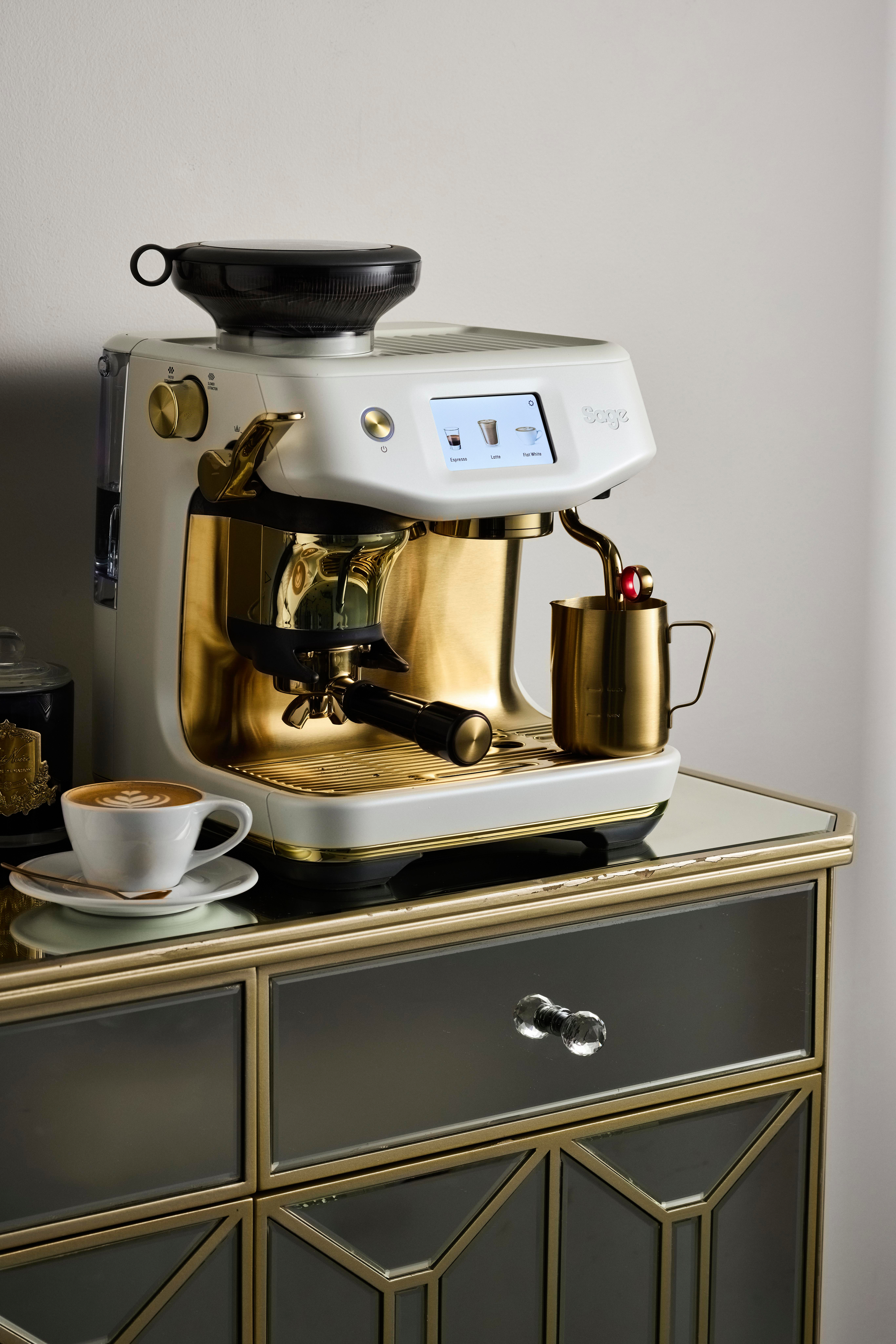 Goldene und weiße Espressomaschine mit einer Tasse Kaffee auf einem verspiegelten Tisch. Eine Milchaufschäumkanne steht unter dem Auslauf.
