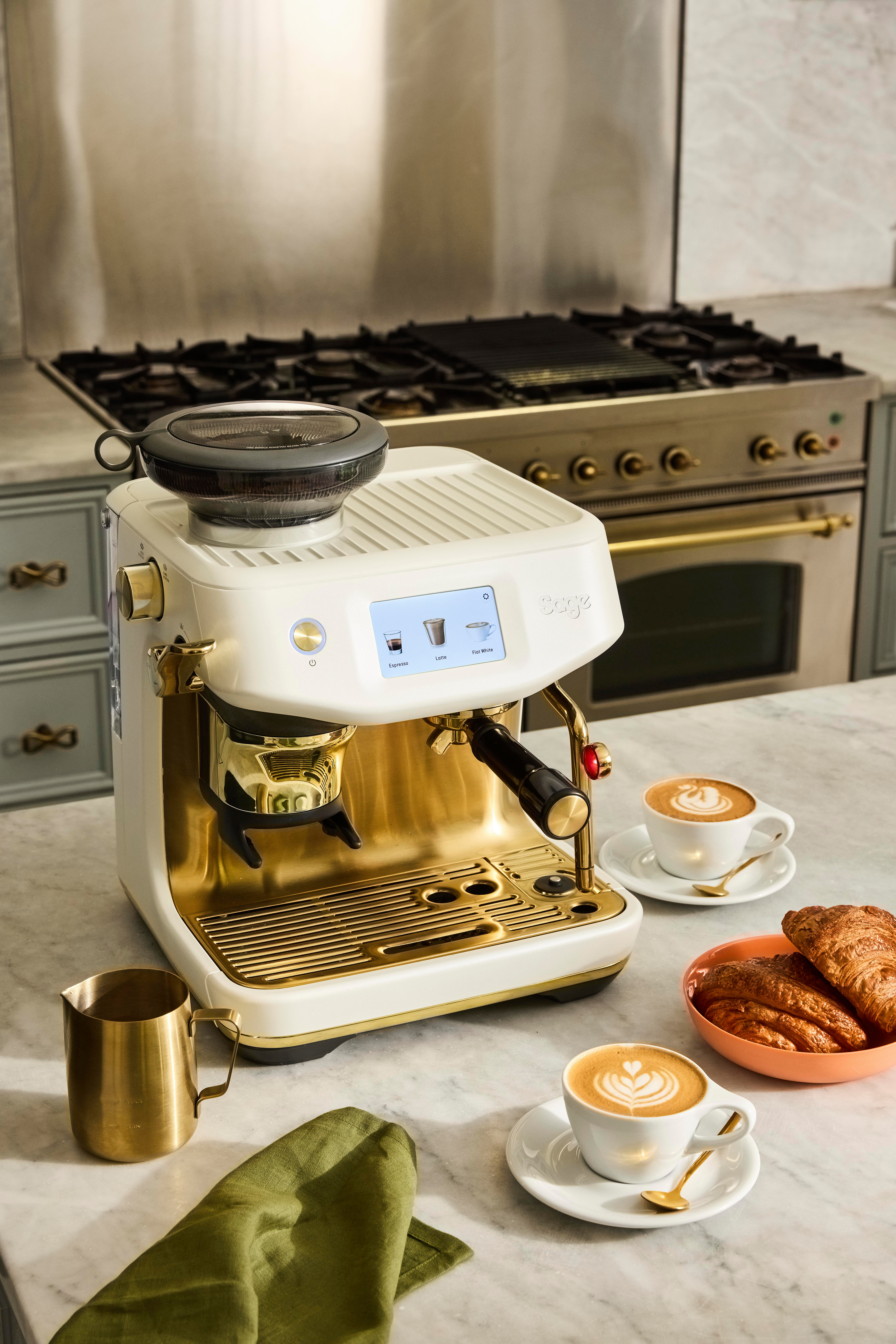 Espressomaschine mit Kaffeetassen, Croissants und einem Krug auf einer Marmorarbeitsplatte. Im Hintergrund ein Herd.