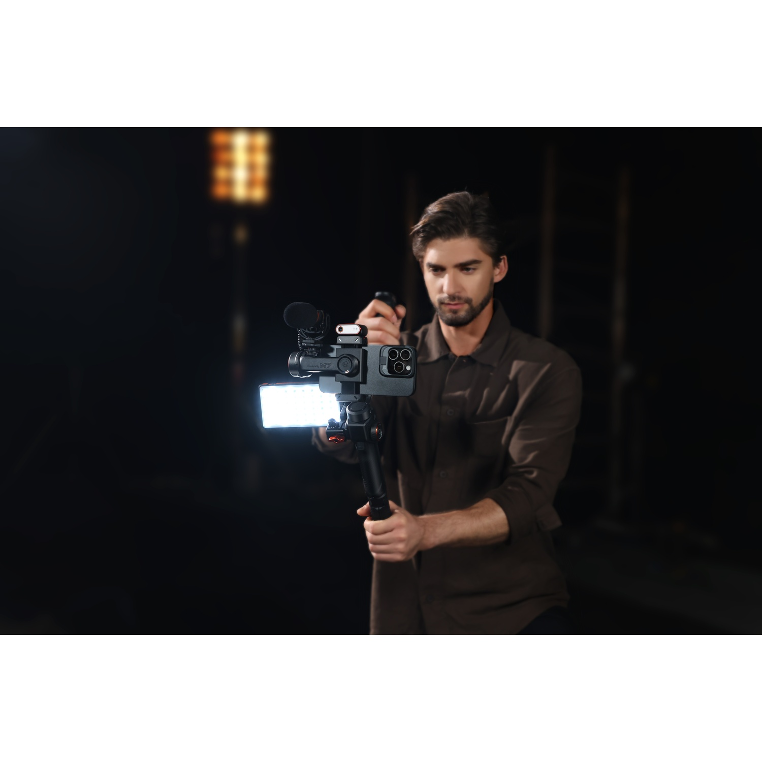 Un uomo tiene un rig per fotocamera con telefono, microfono e luce. Indossa una camicia marrone su sfondo scuro, riprendendo.