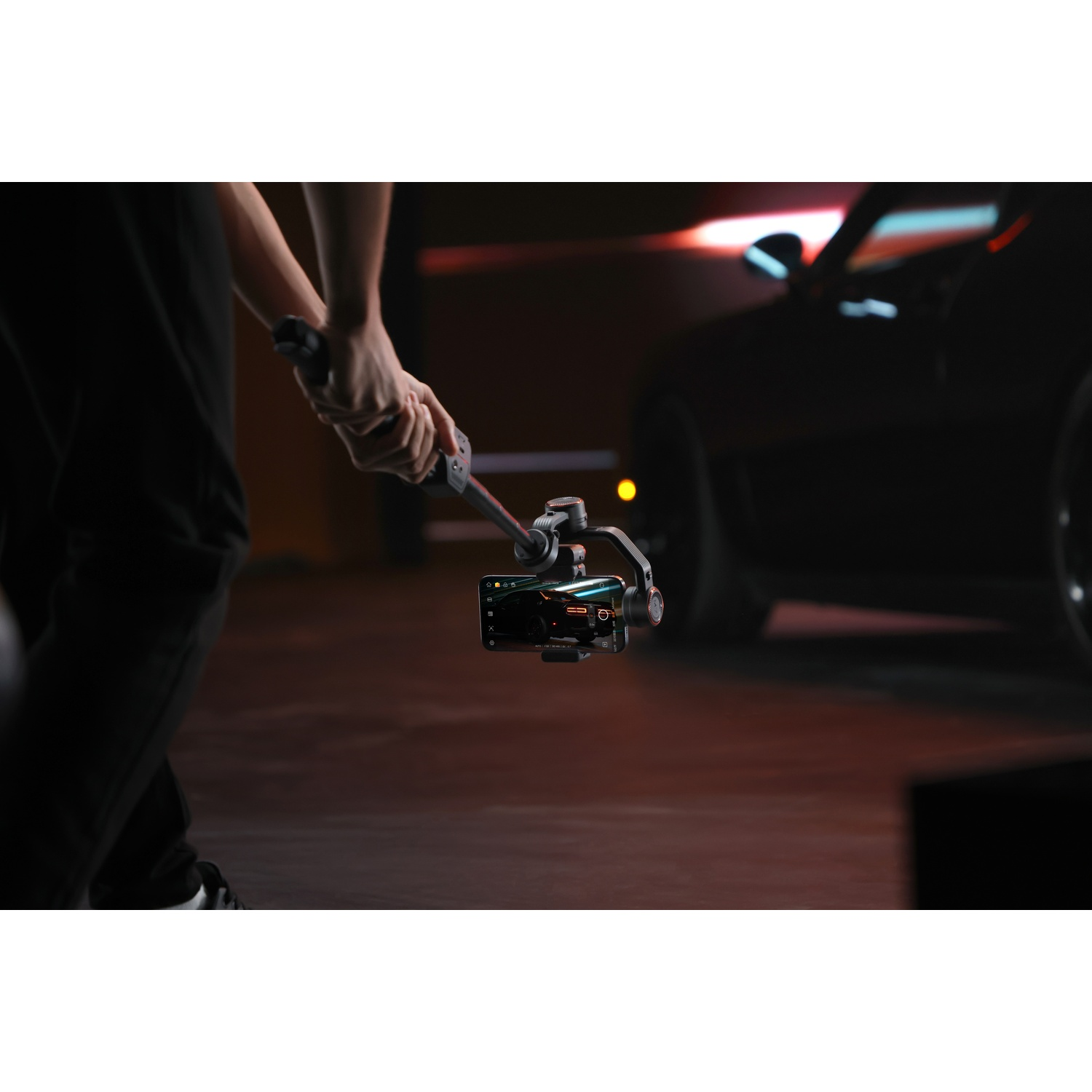 Una persona tiene in mano un gimbal con un telefono, riprendendo un'auto nera in un garage buio. La scena è debolmente illuminata con sfumature rosse.
