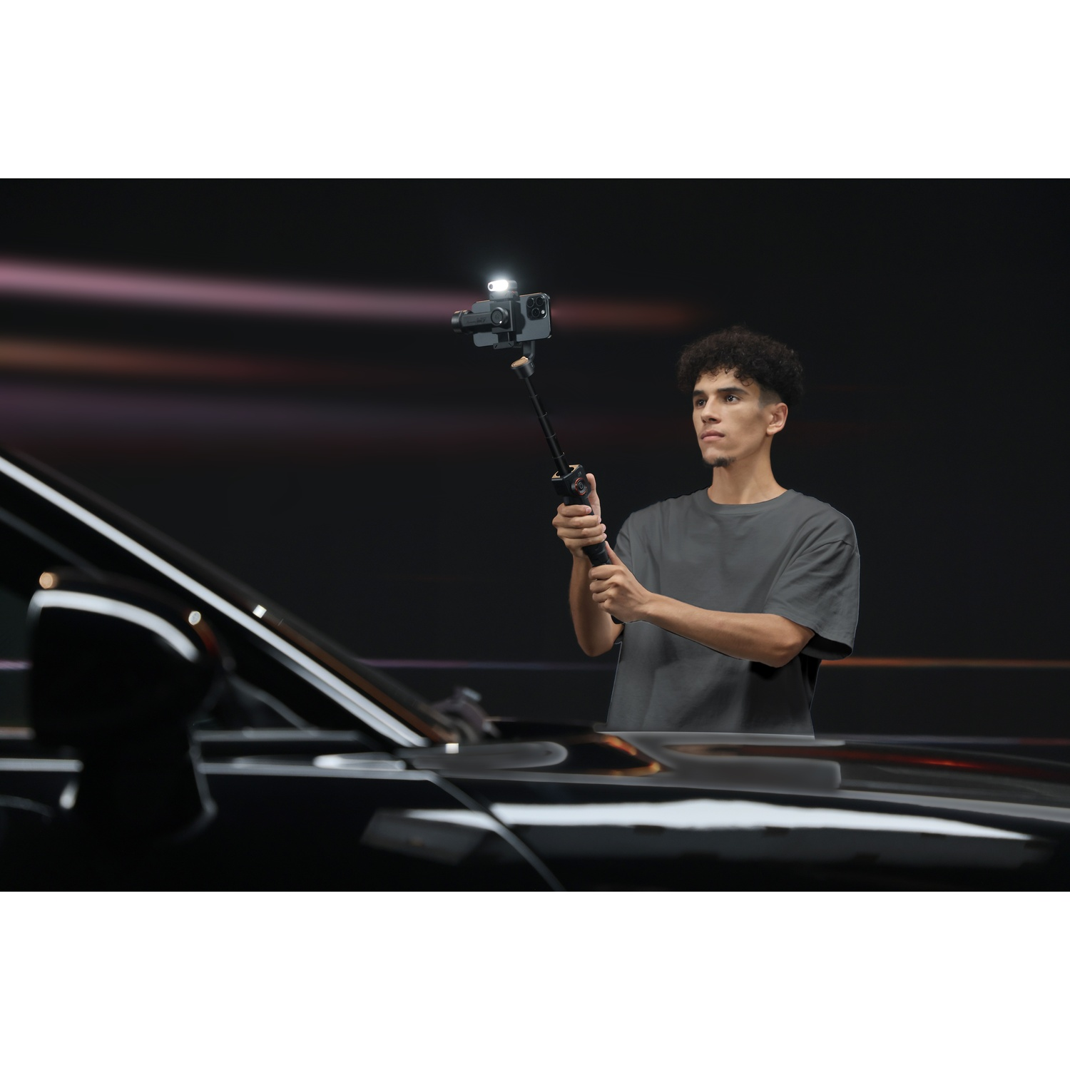 Un uomo tiene un telefono su un selfie stick. Si trova vicino a un'auto nera, con sfondo scuro e luce.