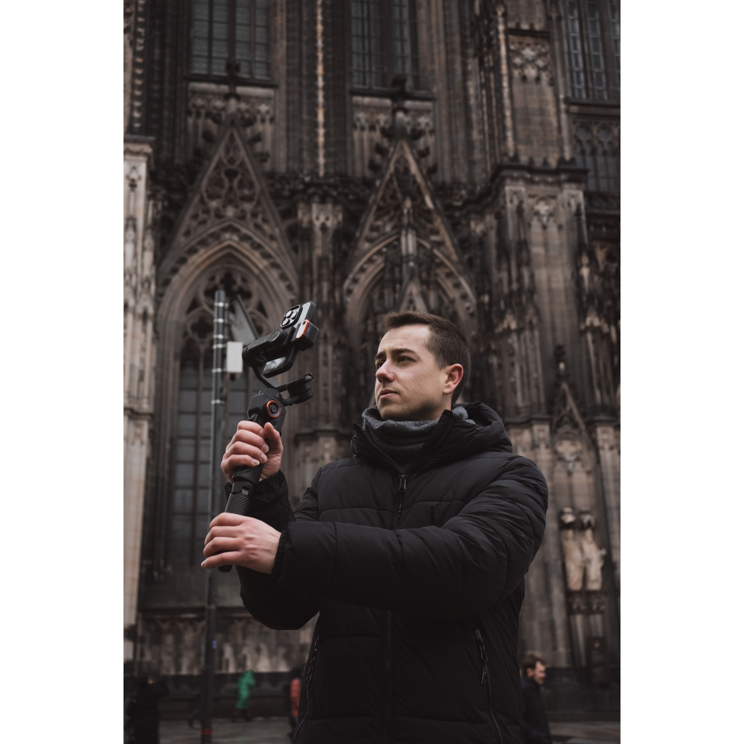 Uomo con stabilizzatore per fotocamera di fronte a una cattedrale. Indossa una giacca nera. La cattedrale è gotica.
