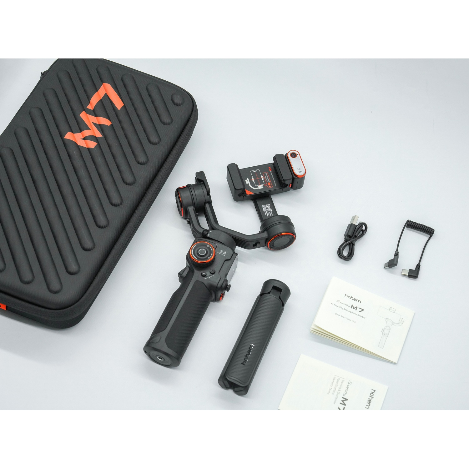 Un gimbal Hohem M7 nero con accessori: custodia, manuali, cavi e un supporto, tutto su una superficie bianca. La custodia ha un 'M7' arancione.