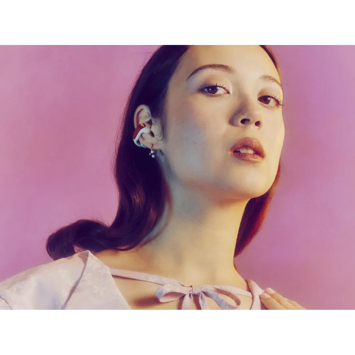 A woman with dark hair and a colorful ear cuff. She wears a light pink dress, with a bow. Pink background with light.