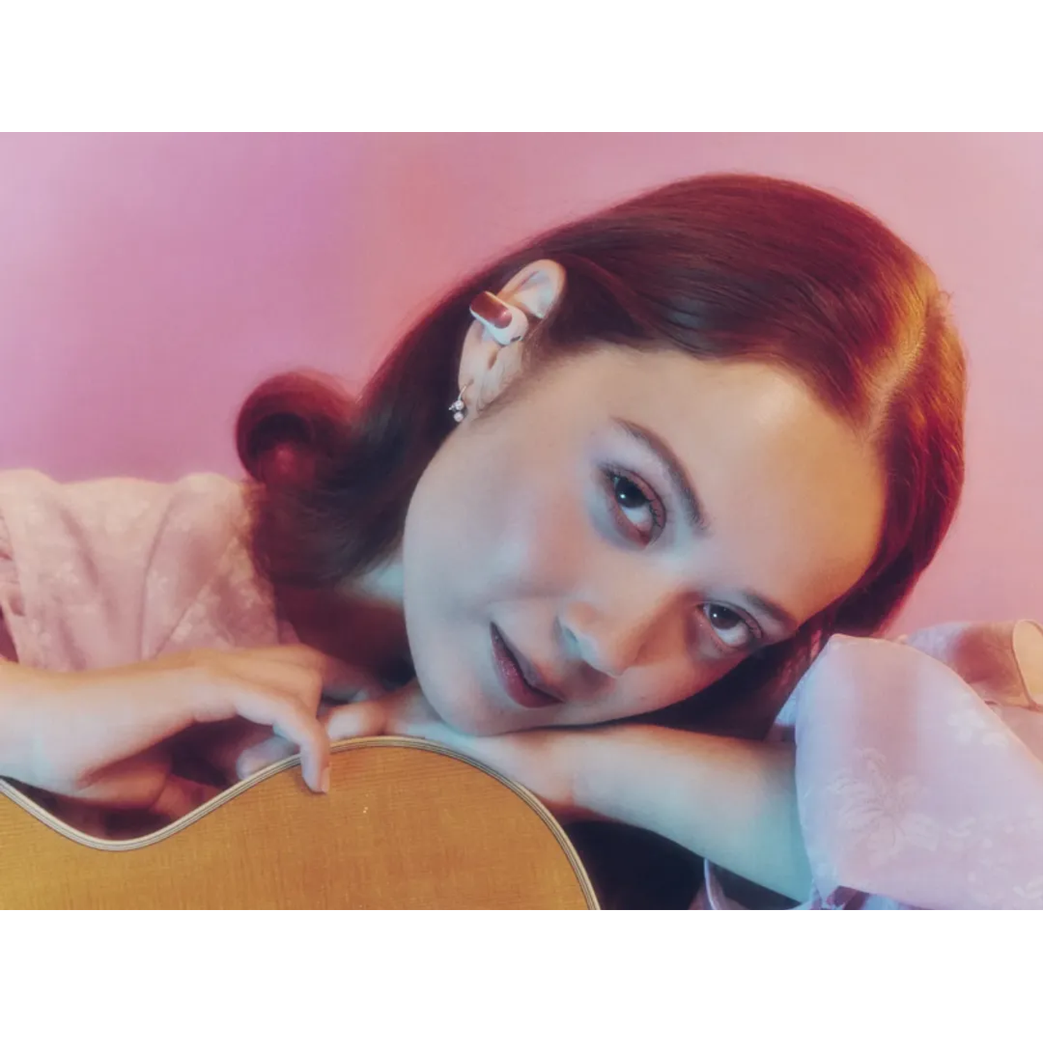 A woman with red hair rests on a guitar. She wears a pink dress and has an earring. The background is pink and blue.