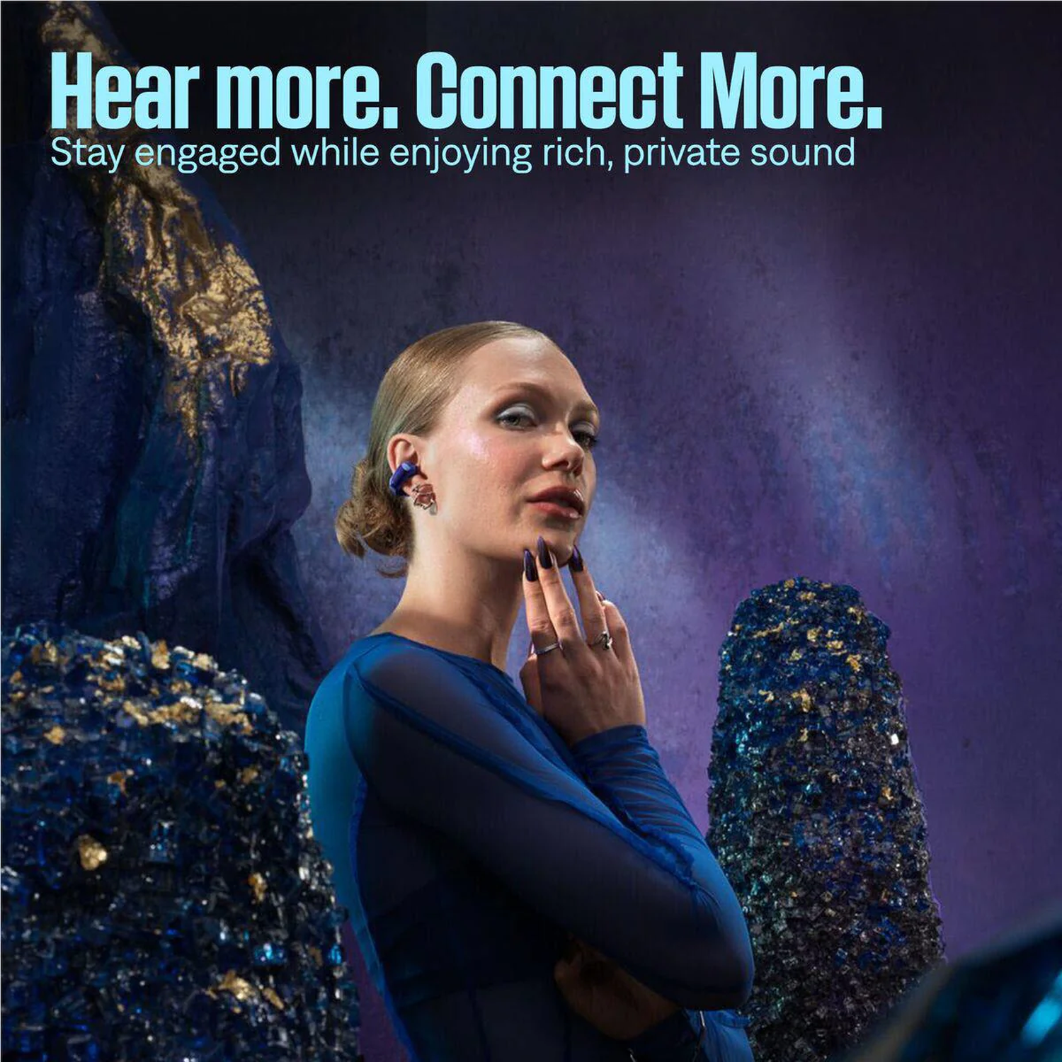Vrouw met blauwe oortelefoons en jurk raakt haar gezicht aan. Donkerblauwe en gouden kristalformaties op de achtergrond. Tekst: Hear more. Connect More.