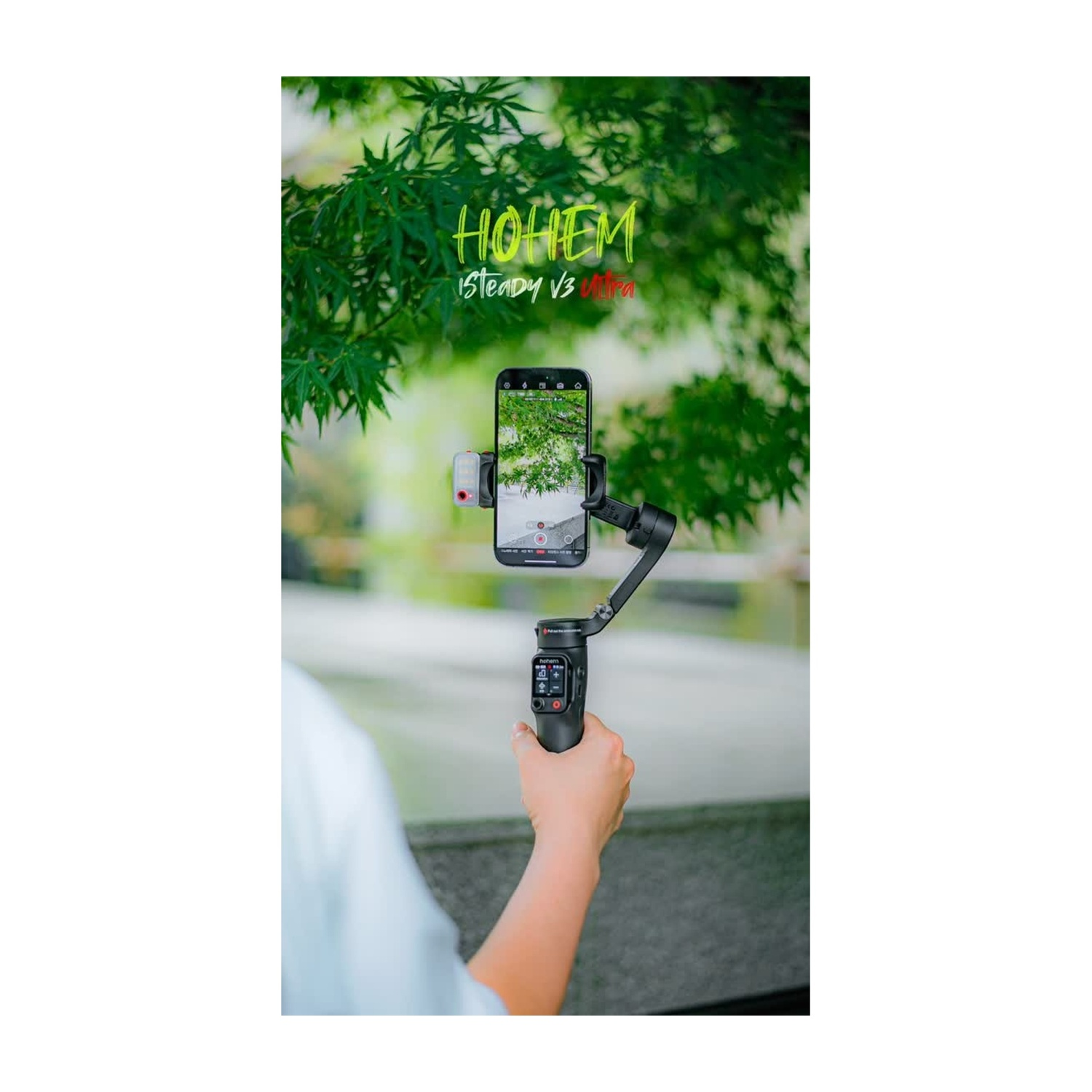 Una persona tiene in mano un telefono su un gimbal, che filma un albero verde. Lo sfondo è una scena sfocata di un parco con fogliame verde e acqua.