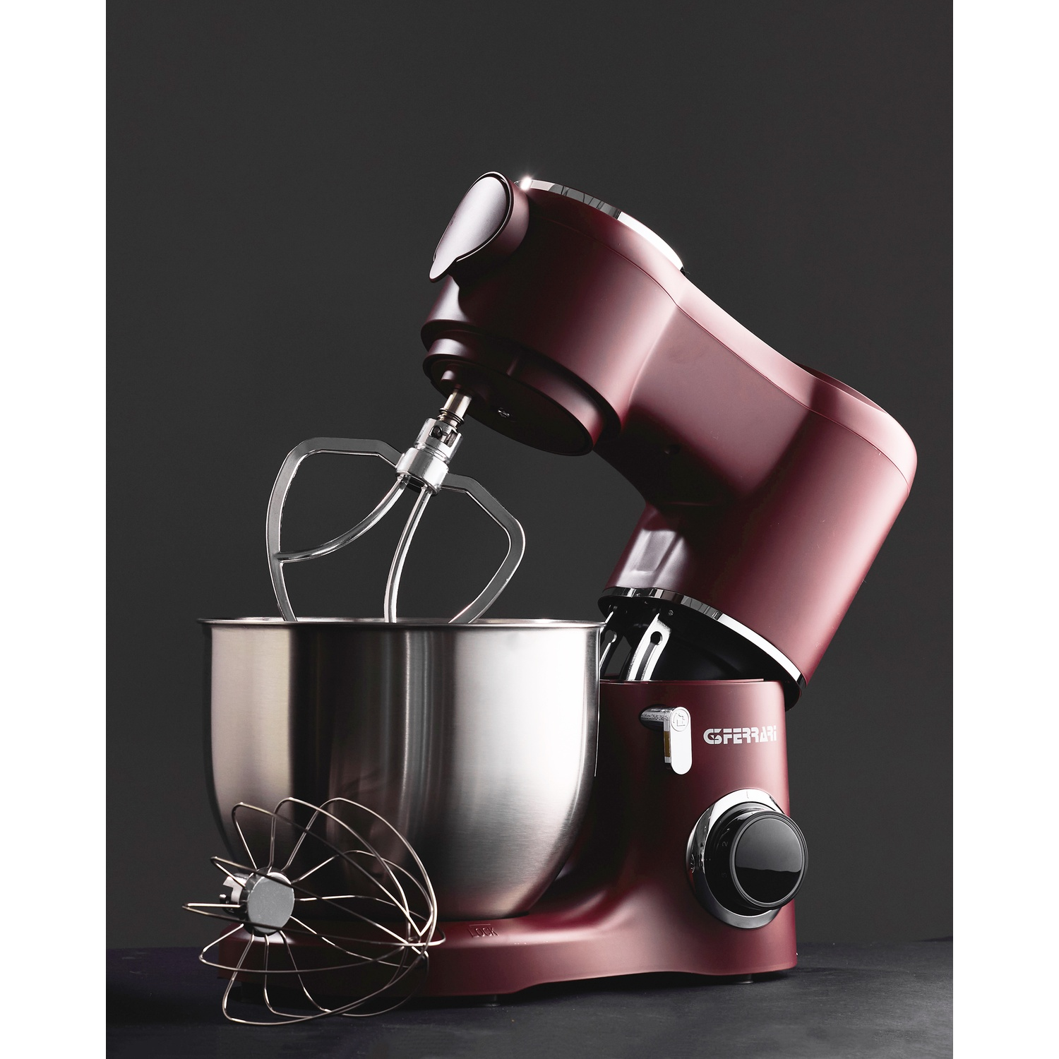 Un mixer da cucina bordeaux con ciotola e accessori in acciaio inox. Una frusta è a sinistra. Lo sfondo è grigio scuro.