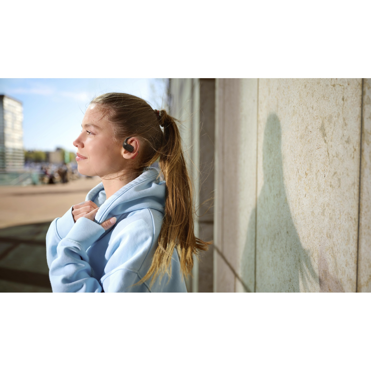 Una giovane donna con una felpa blu e un auricolare guarda altrove. Si trova vicino a un muro strutturato in una giornata di sole.