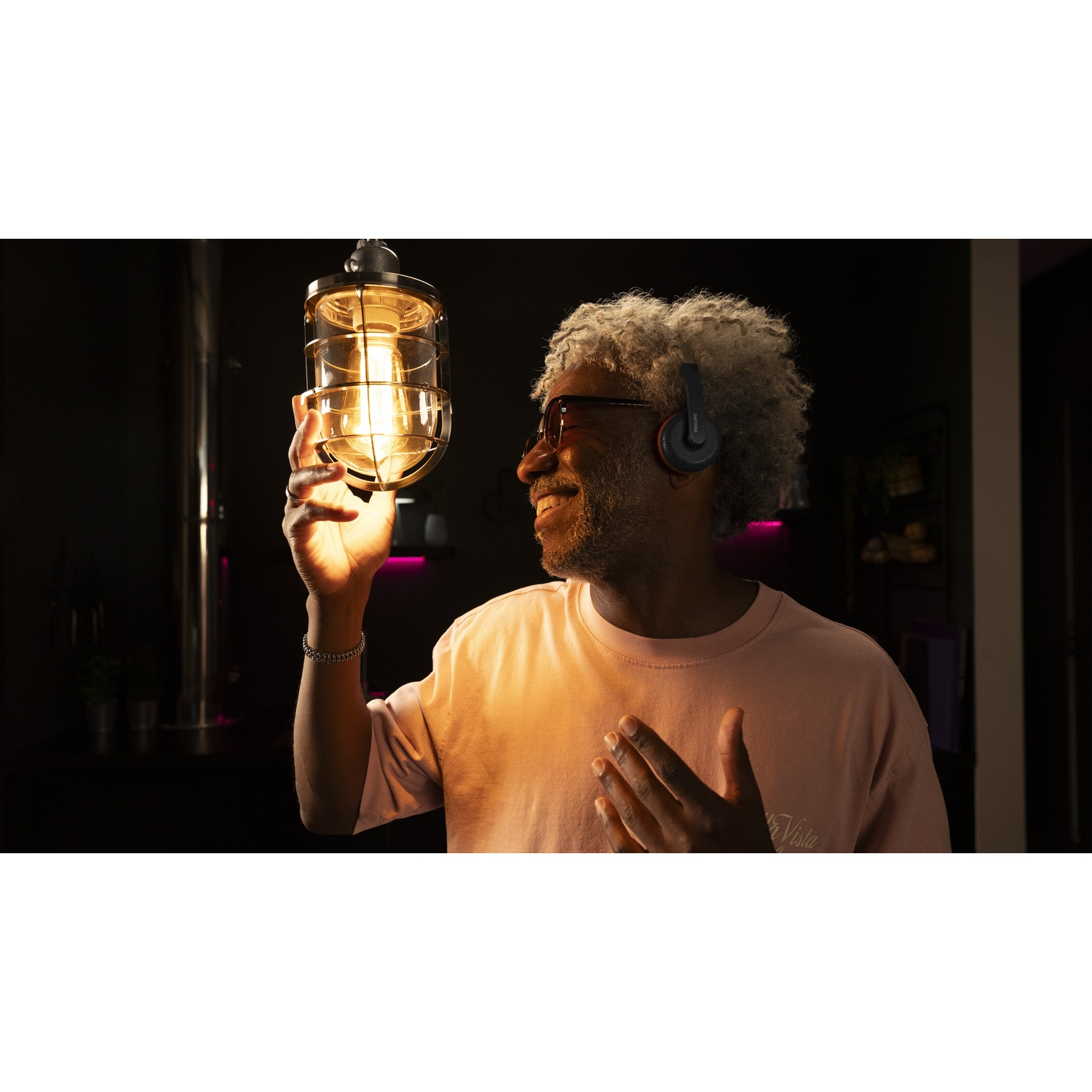 Un uomo con i capelli grigi e occhiali da sole esamina una lampada accesa e ingabbiata. Indossa una camicia rosa e delle cuffie, sorridendo. Sfondo scuro.