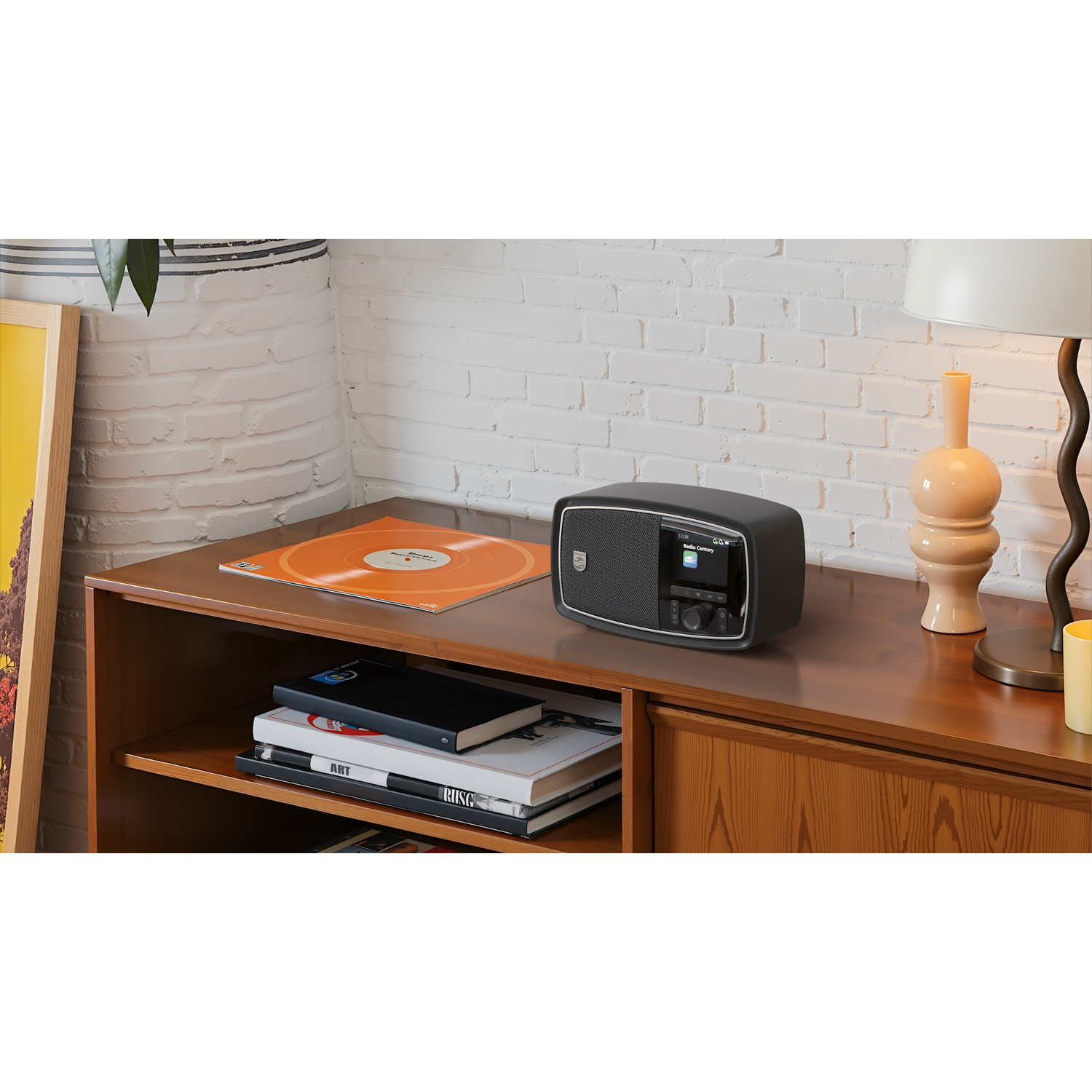 Una radio nera è su un mobile in legno, vicino a un disco e libri. Una lampada e un muro di mattoni sono sullo sfondo.
