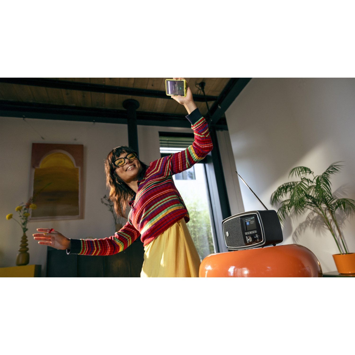 Donna con maglione colorato fa un selfie. Una radio è su uno sgabello arancione. Piante e un dipinto sullo sfondo.