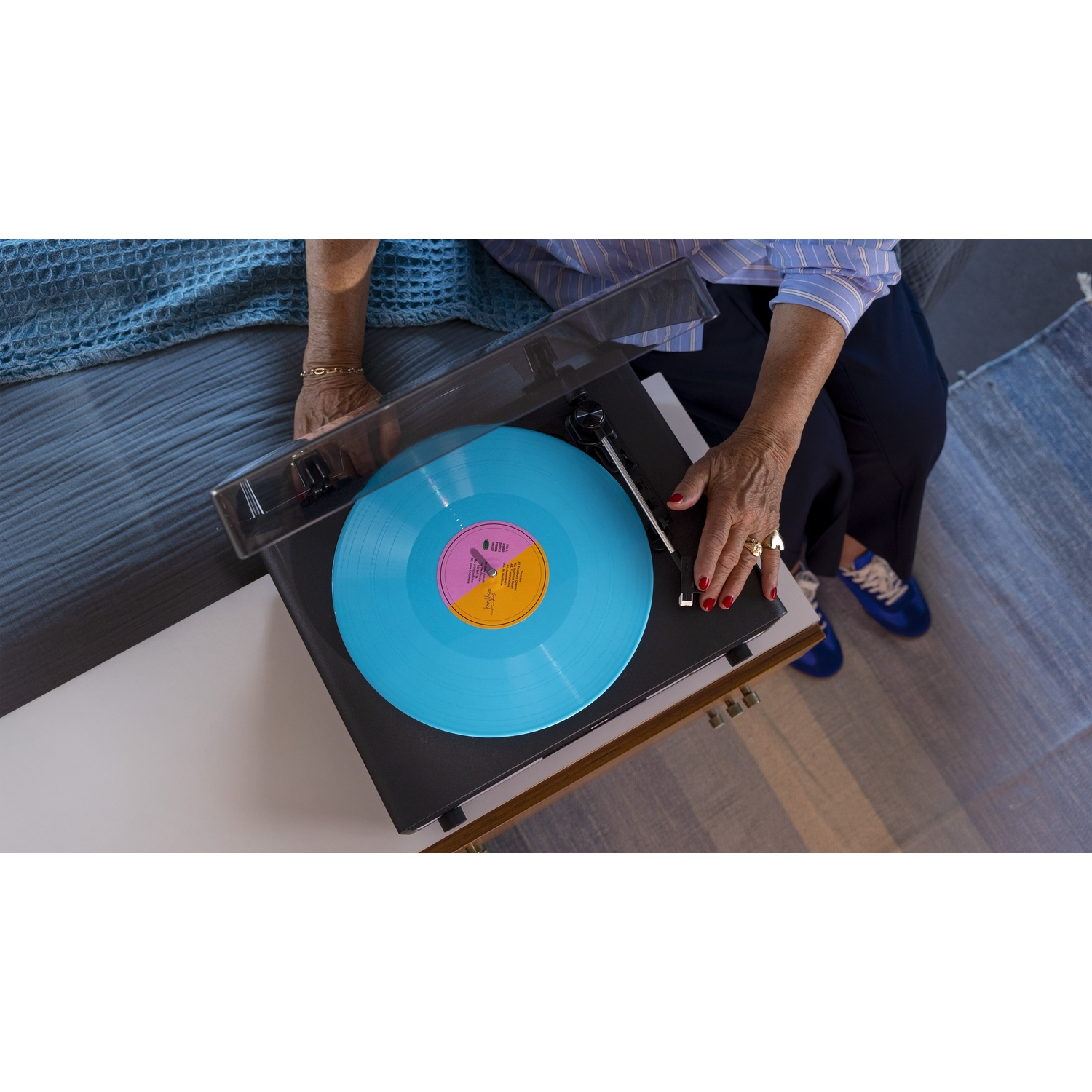 Mani con unghie rosse su un giradischi con un vinile blu. Una persona con scarpe da ginnastica blu è vicina. La scena è in interni.