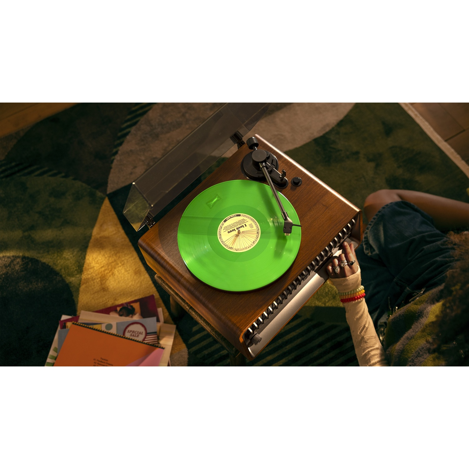 Una persona giace vicino a un giradischi con un disco in vinile verde. Braccialetti e anelli colorati adornano la mano della persona. Libri sono accanto al lettore.