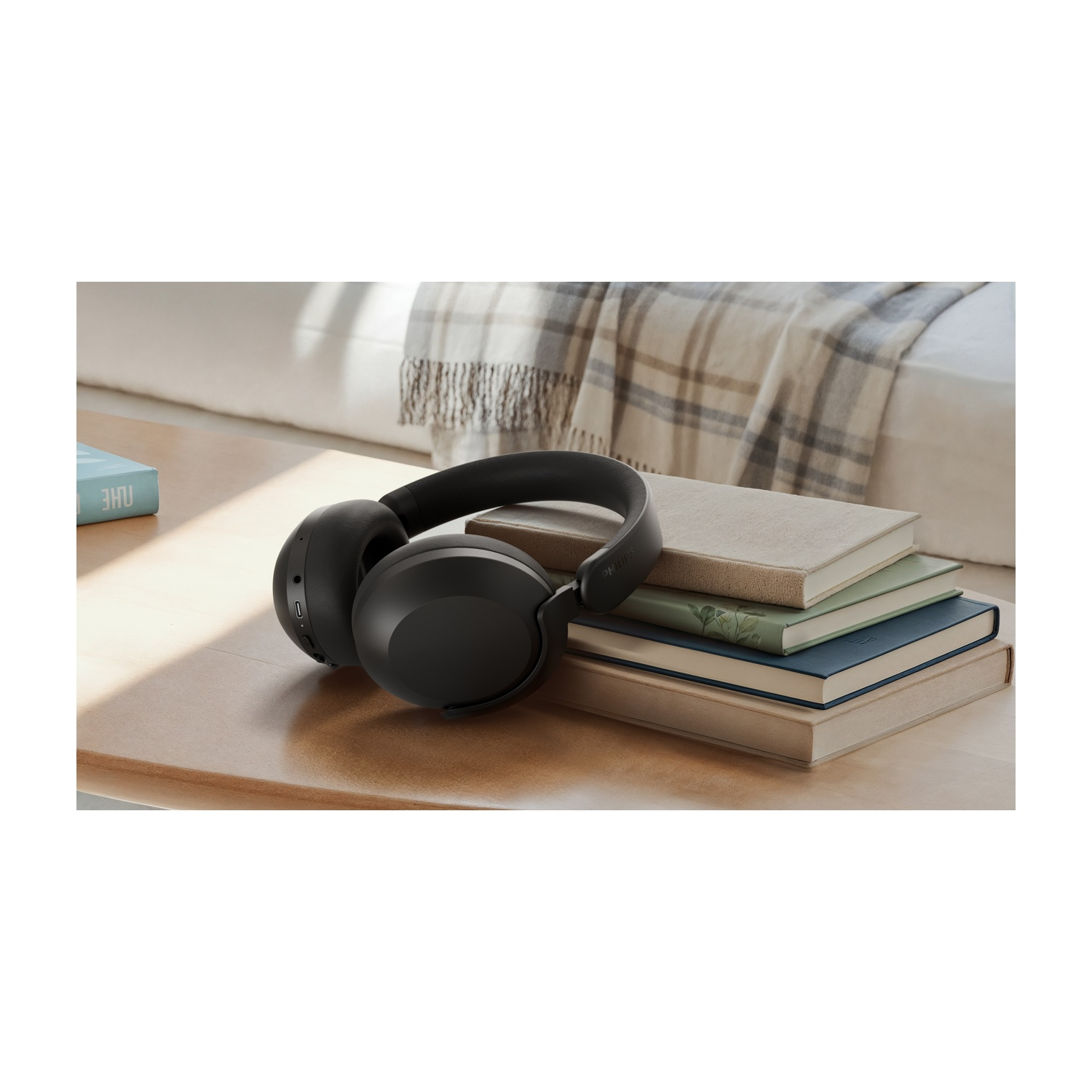 Cuffie nere su un tavolo di legno, accanto a una pila di libri. Una coperta e un letto sullo sfondo.