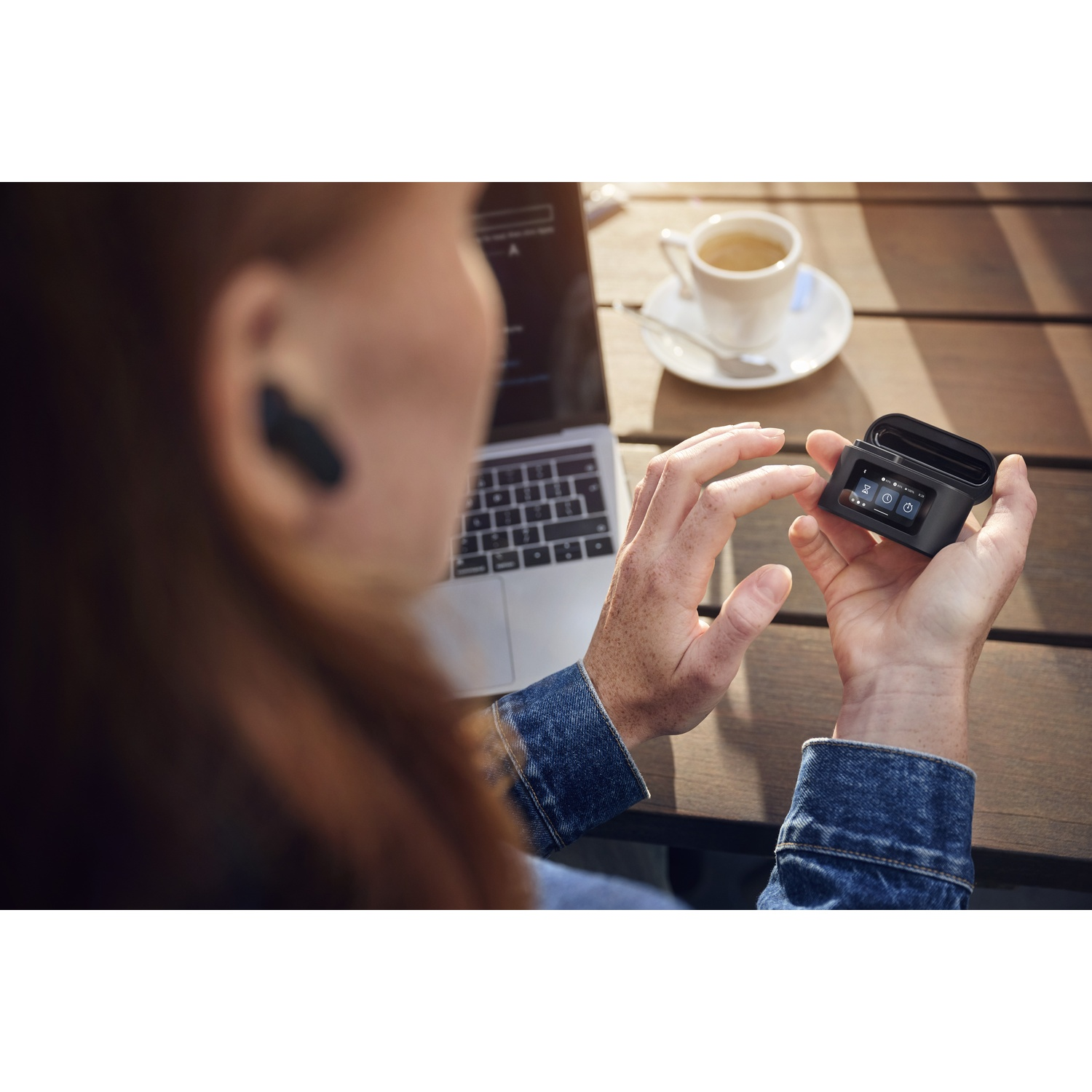 Una persona con un auricolare tocca un dispositivo nero. Un laptop e una tazza di caffè sono su un tavolo di legno.