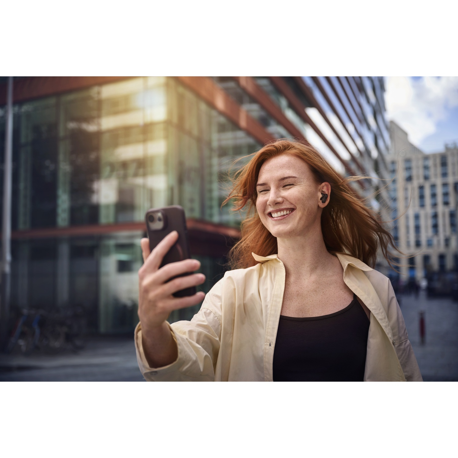 Una donna sorridente con i capelli rossi si fa un selfie con un telefono. Indossa un top nero e una giacca color crema, con edifici sullo sfondo.