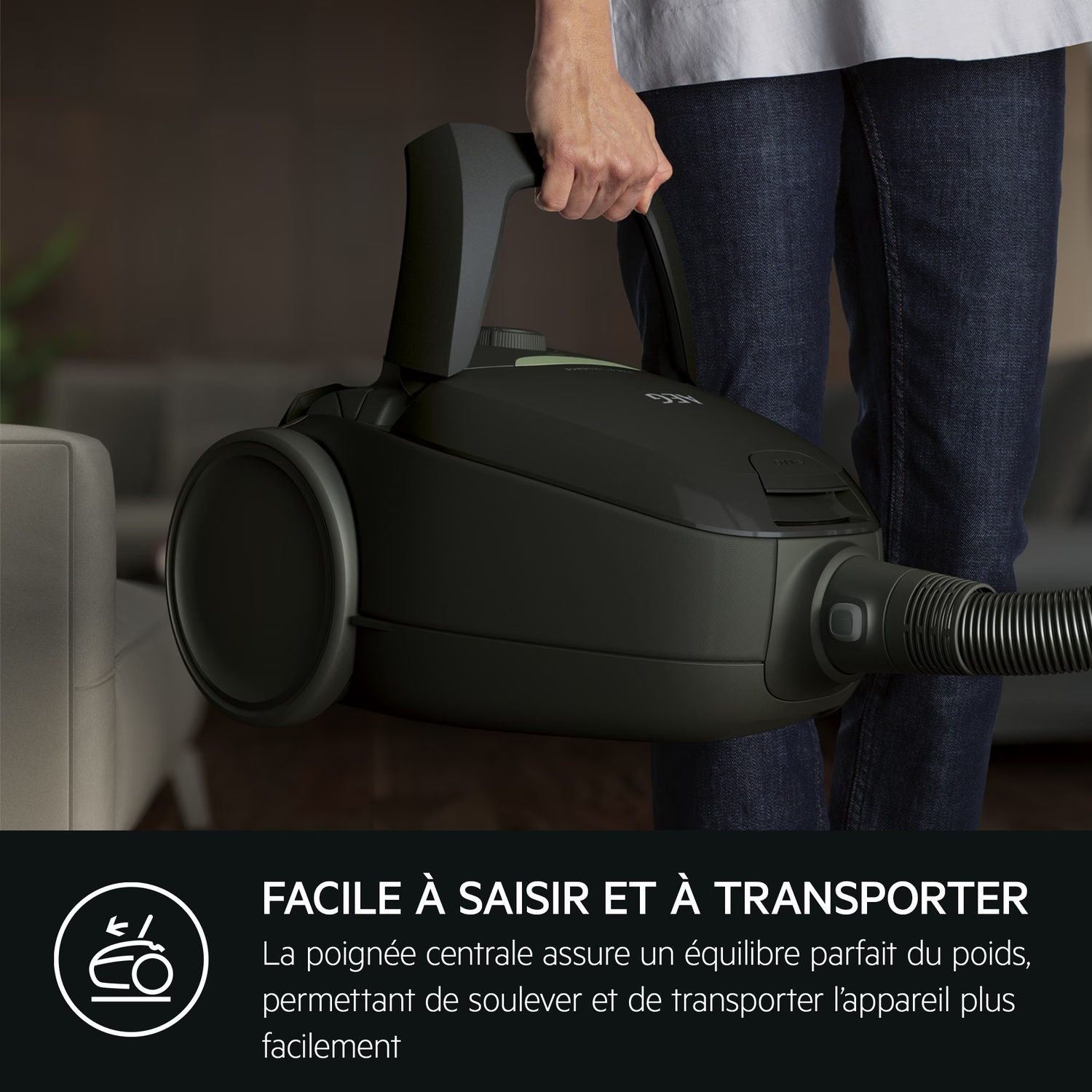 Une personne tient un aspirateur noir. L'aspirateur est soulevé. La personne porte un jean et une chemise blanche.