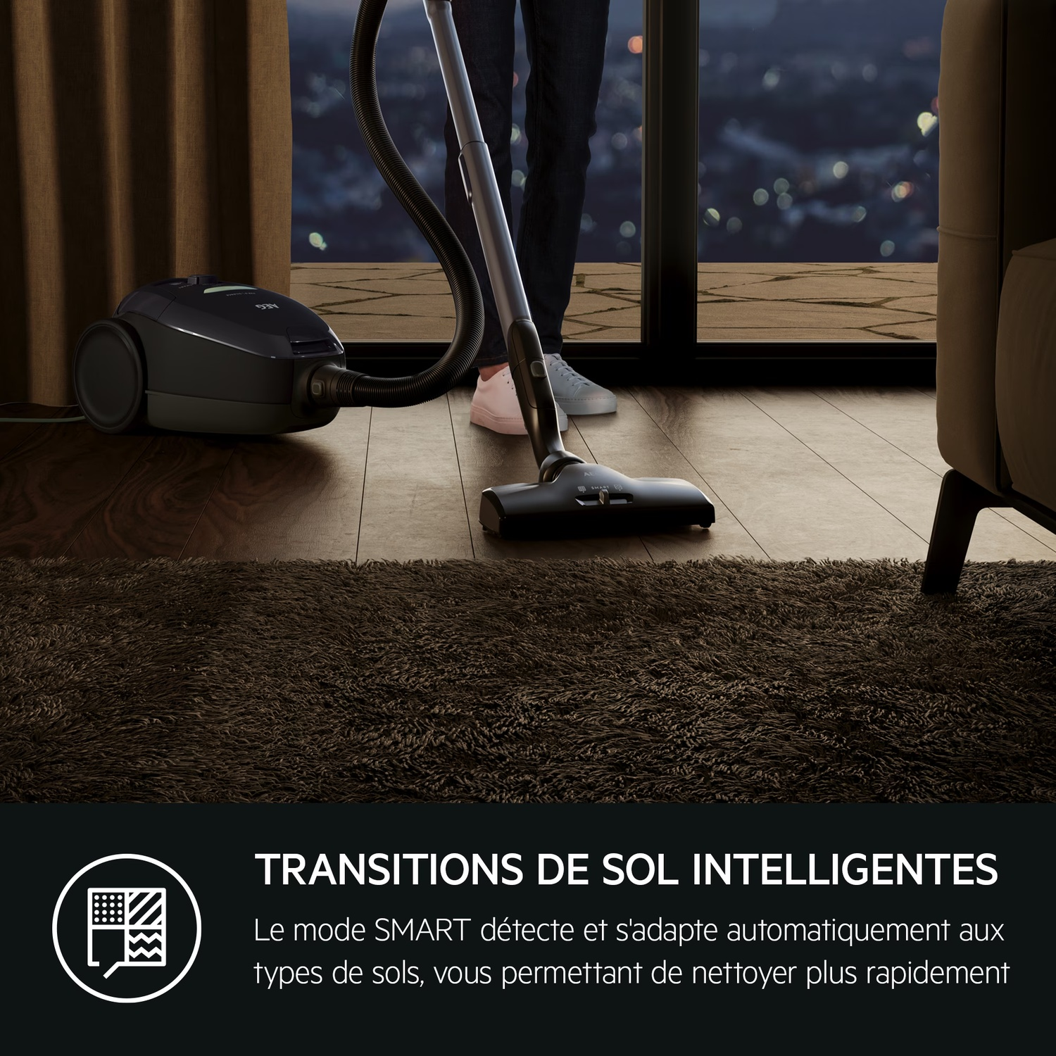 Une personne passe l'aspirateur sur un tapis brun foncé avec un aspirateur noir. L'arrière-plan montre une fenêtre avec des lumières de la ville.