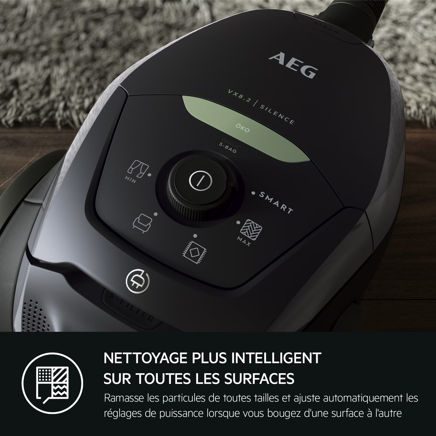 Un aspirateur AEG noir, avec des boutons de commande et un cadran d'alimentation. L'aspirateur est posé sur un sol en bois, avec un tapis en arrière-plan.