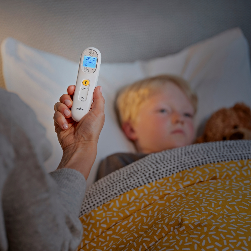 Eine Hand hält ein Thermometer (36,5°C) neben einem Kind im Bett. Ein Teddybär sitzt neben dem Kind unter einer gelben Decke.