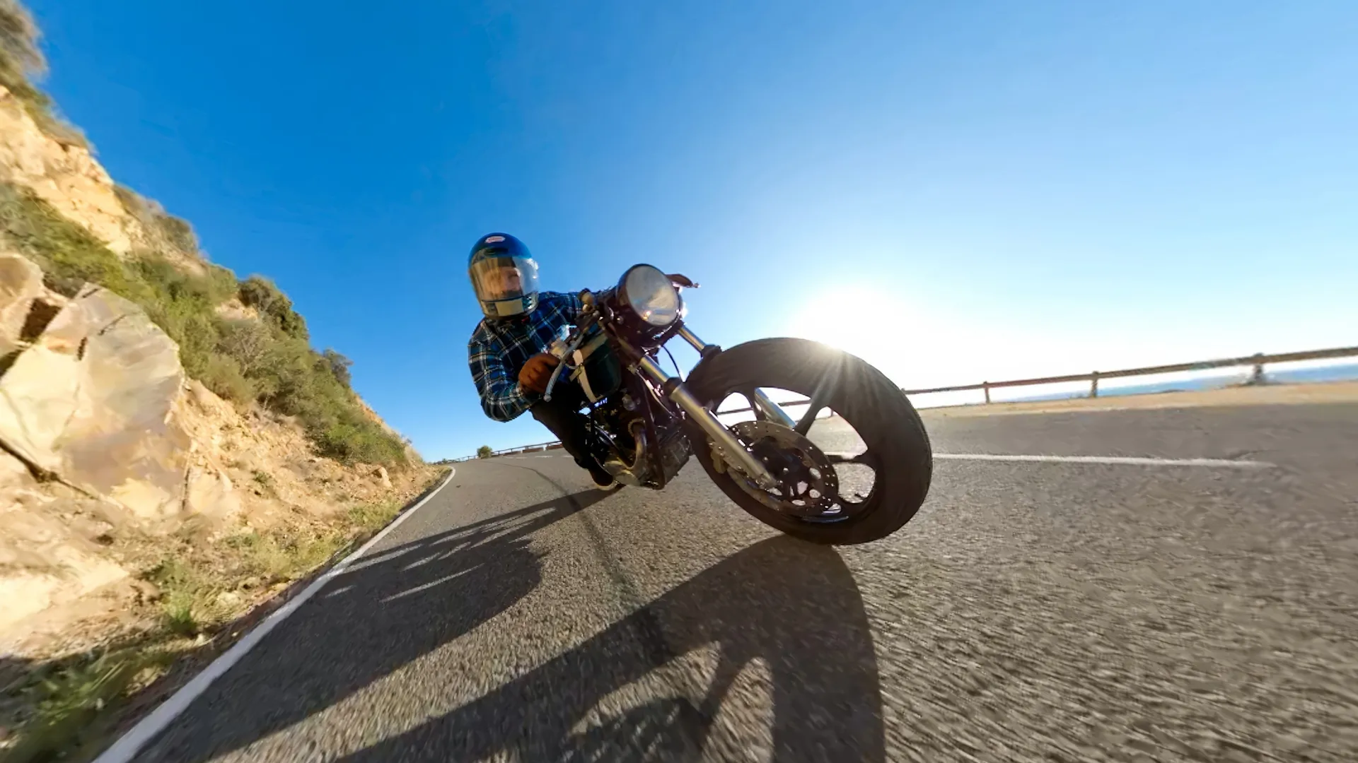 Un motocycliste s'incline dans un virage sur une route. Ciel bleu, montagnes et mer en arrière-plan. Le soleil brille vivement.