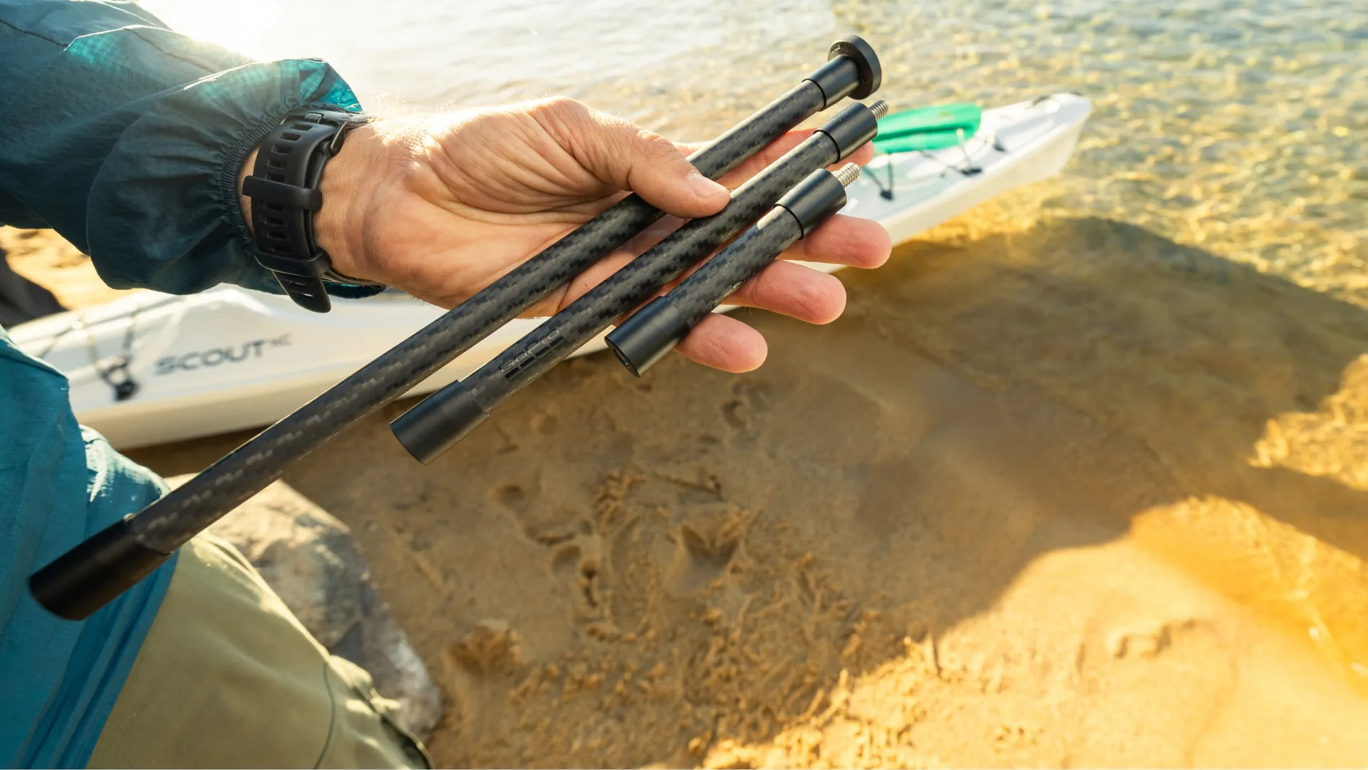 Une main tient trois poteaux noirs près d'un kayak sur une plage. Une montre est au poignet. L'eau est claire.